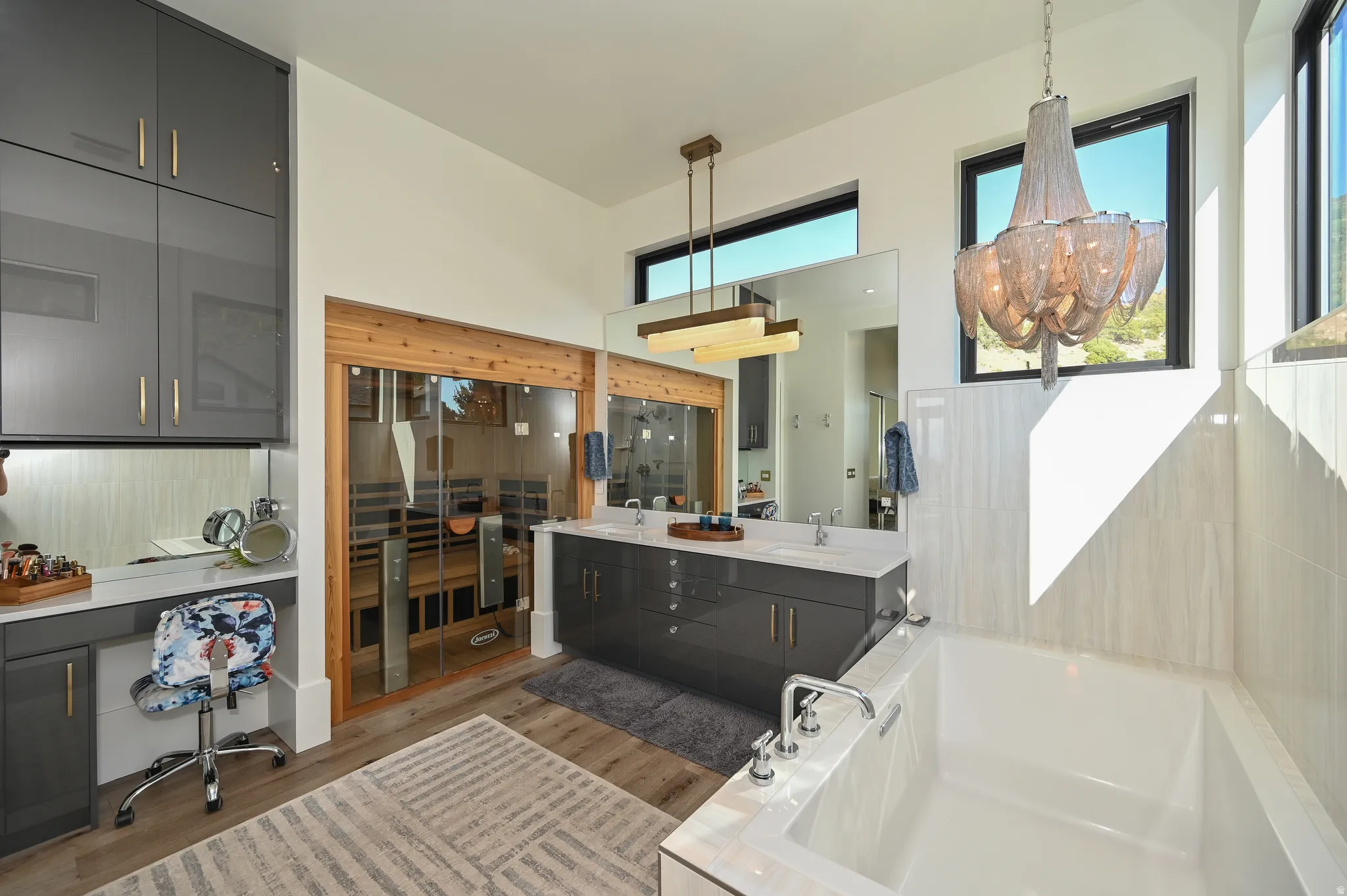 Spacious bathroom featuring a built-in sauna with wood-frame glass doors, a freestanding soaking tub, dual vanity with quartz countertop, a dedicated vanity area with storage, and high ceilings with upper windows and contemporary light fixtures