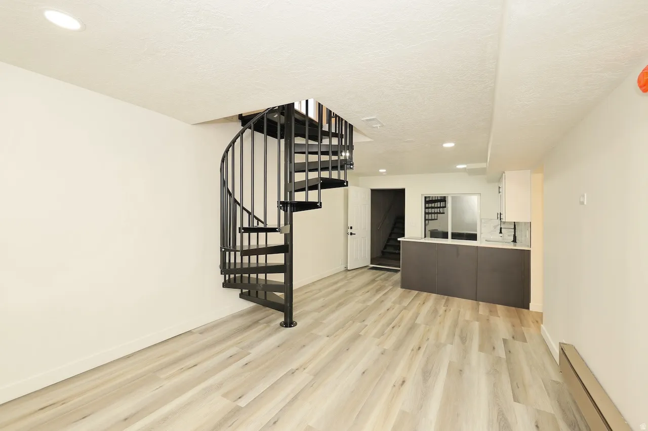 Open-concept living area featuring a black metal spiral staircase, light wood-finish flooring, recessed lighting, white walls, and a built-in counter with white upper cabinetry