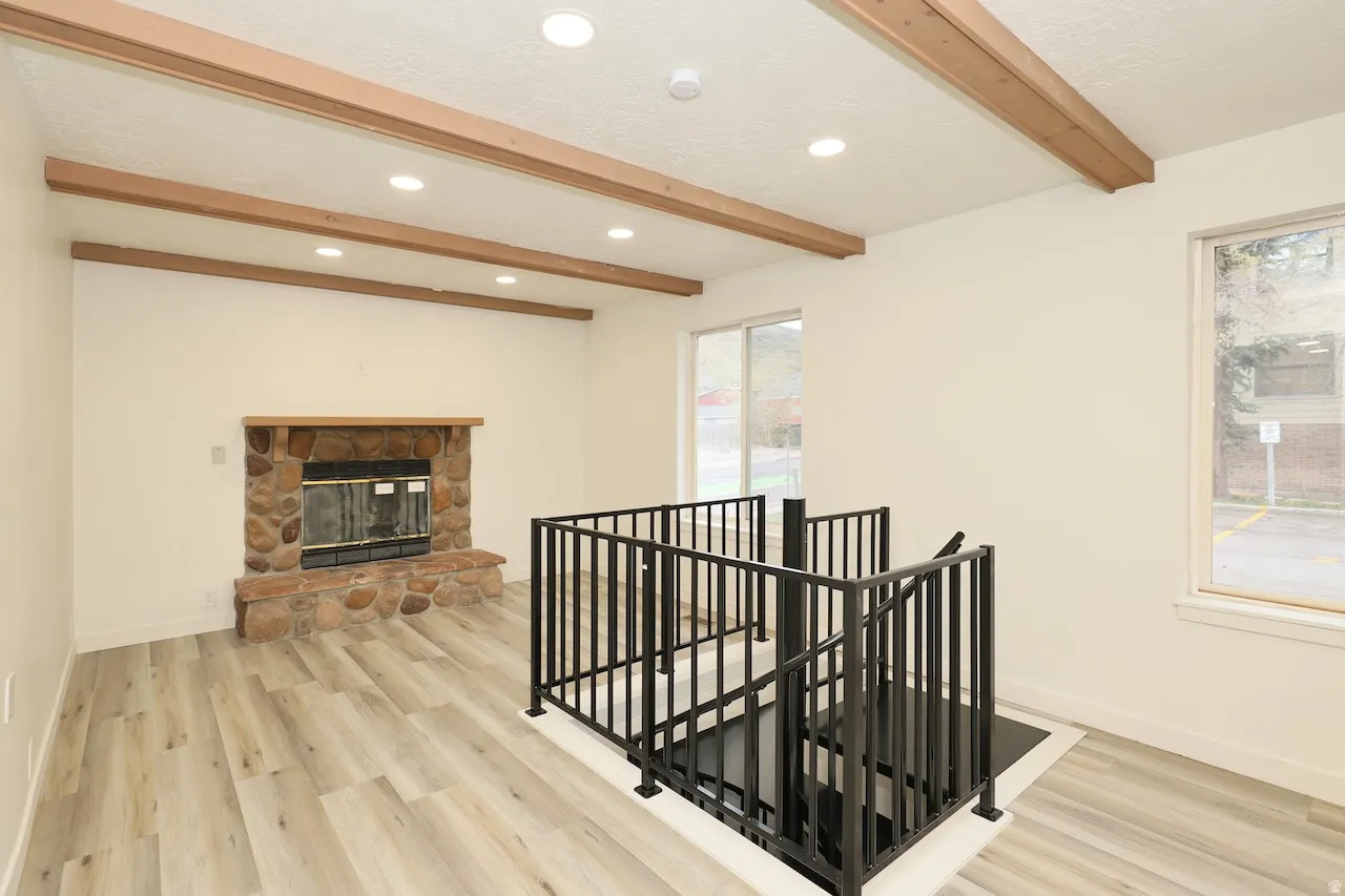 Living area featuring wood-finish flooring, a stone fireplace with a wood mantel, exposed ceiling beams, recessed lighting, and a modern black metal staircase railing