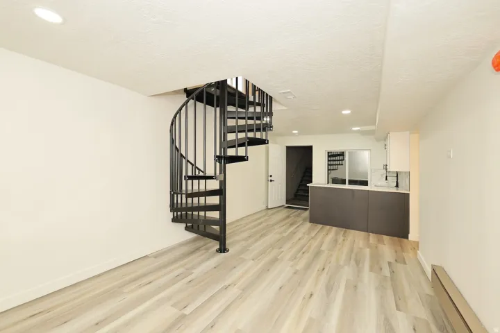 Open-concept living area featuring a black metal spiral staircase, light wood-finish flooring, recessed lighting, white walls, and a built-in counter with white upper cabinetry