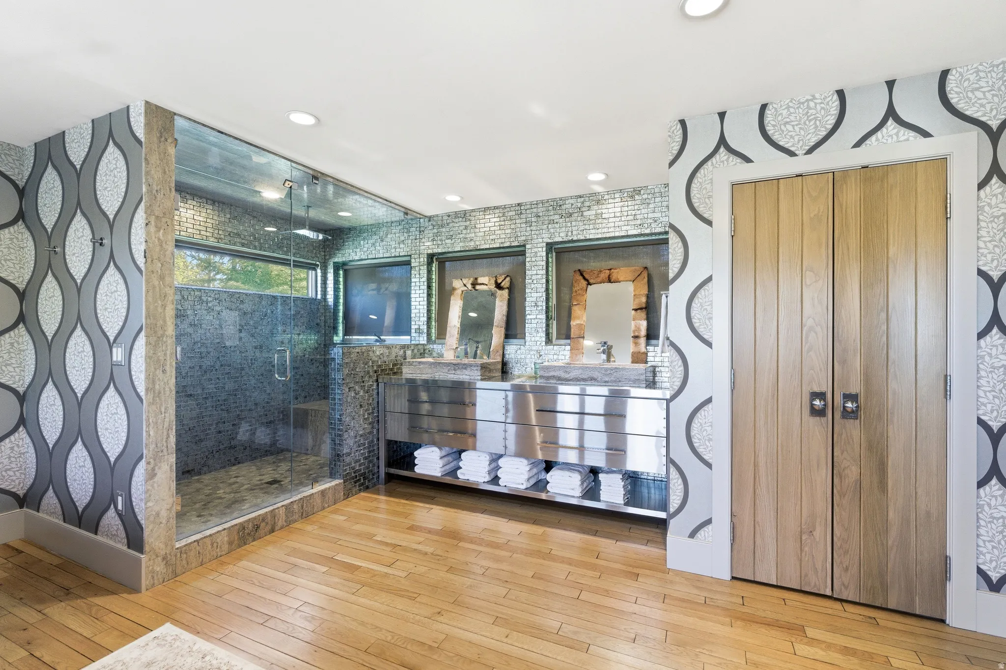 Spacious bathroom featuring a glass-enclosed shower with mosaic tile, double vanity with dual vessel sinks and under-cabinet storage, wood-finish flooring, and natural wood bi-fold doors