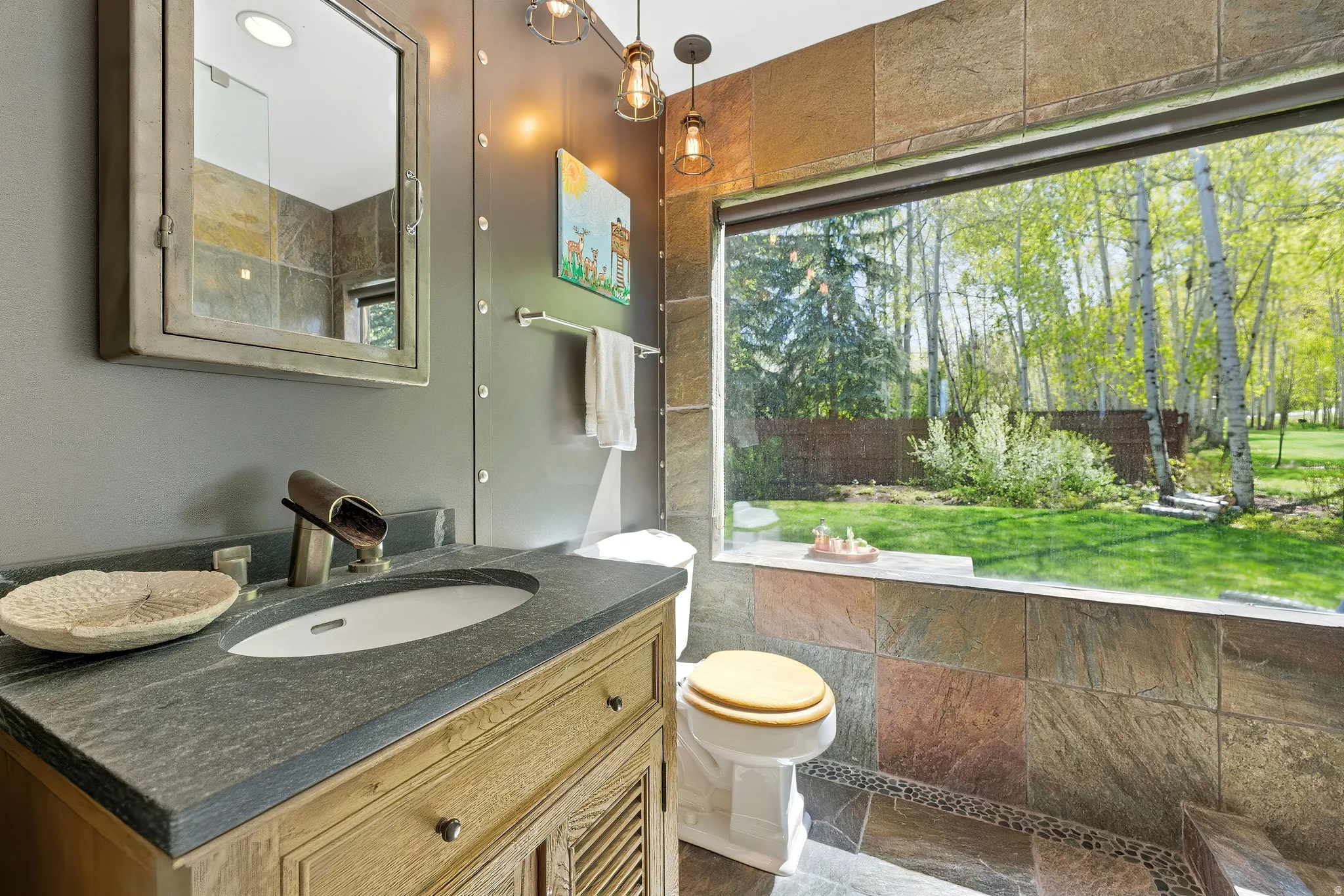 Bathroom featuring an expansive horizontal window, stone tile walls, a stone vanity countertop, a wood-finish vanity cabinet, and industrial-style pendant lighting