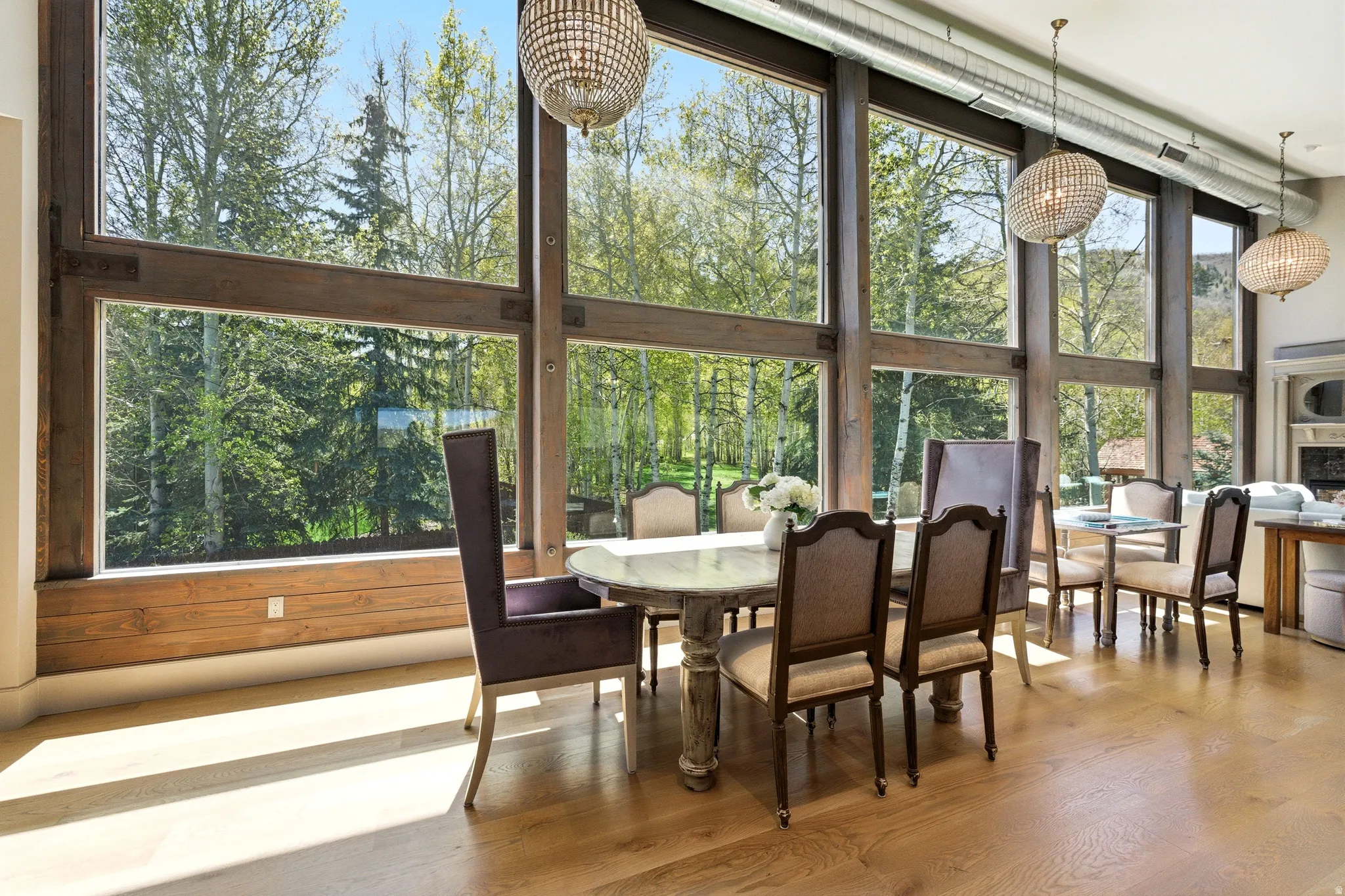 Expansive floor-to-ceiling windows with wood framing, wood-finish flooring, two ornate beaded chandeliers, and exposed metal ductwork