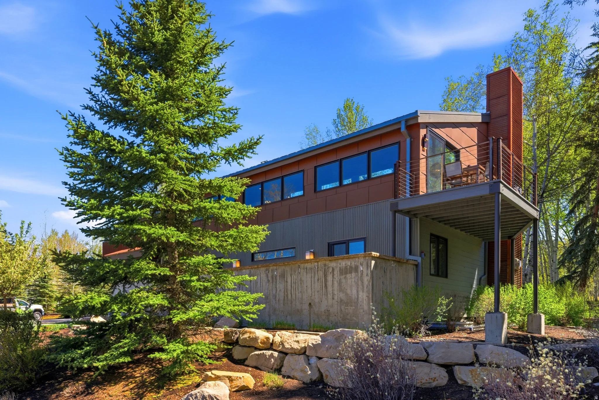 Contemporary architectural design featuring multi-toned horizontal siding, expansive upper-level windows, a cantilevered balcony with metal railing, and a prominent stone retaining wall