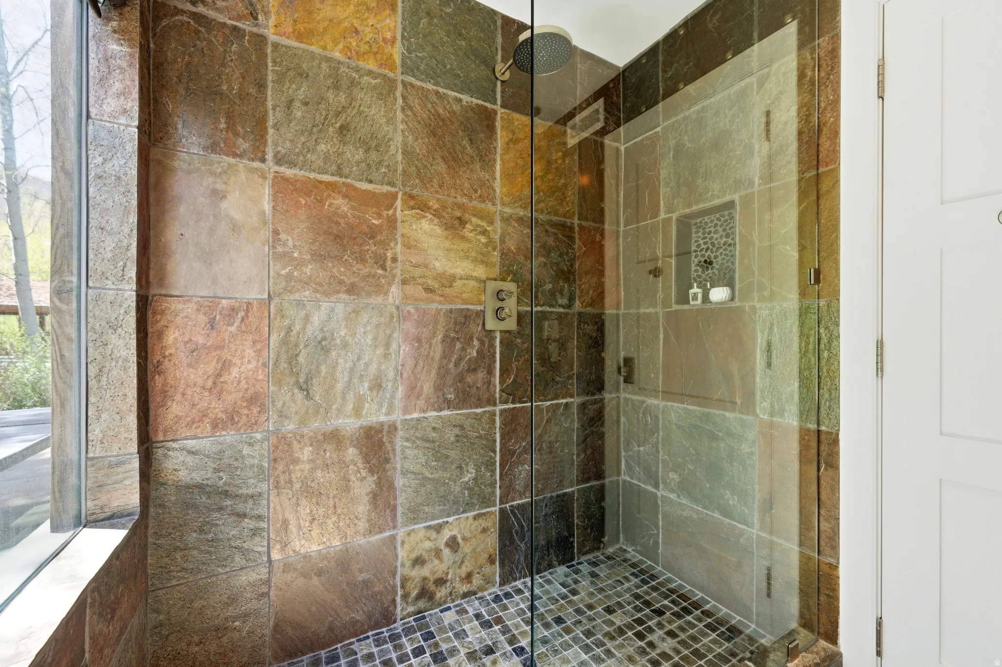 Shower enclosure featuring natural stone wall tiling, a glass-panel door, and a built-in shower niche with mosaic tile accents