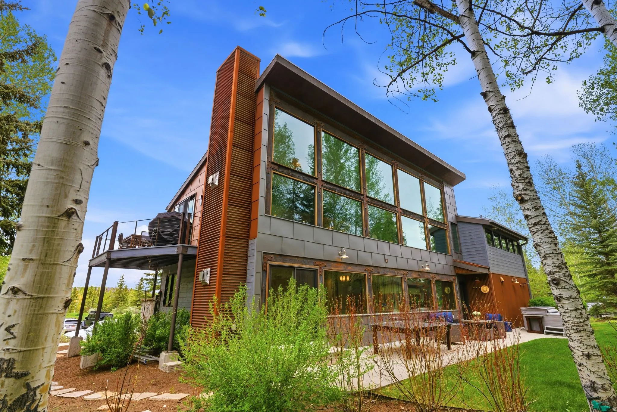 Contemporary property with extensive window fenestration, a sizable upper-level deck with metal railings, a ground-level patio, and a mix of vertical wood-finish siding and horizontal paneling