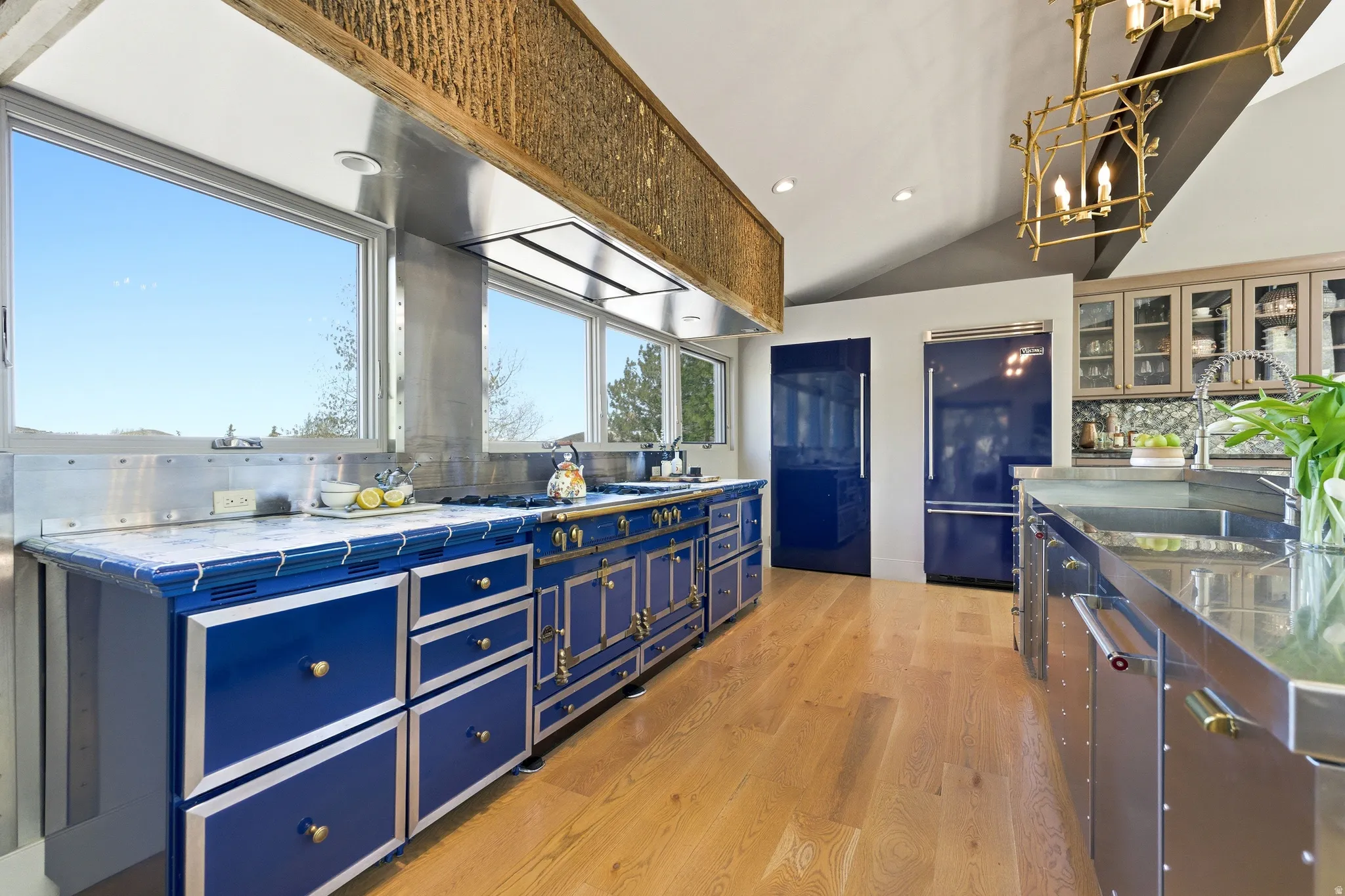 Gourmet kitchen featuring a vibrant blue range with brass hardware, a matching integrated refrigerator and freezer, wood-finish flooring, a stainless steel island with an integrated sink, and an exposed wood beam ceiling