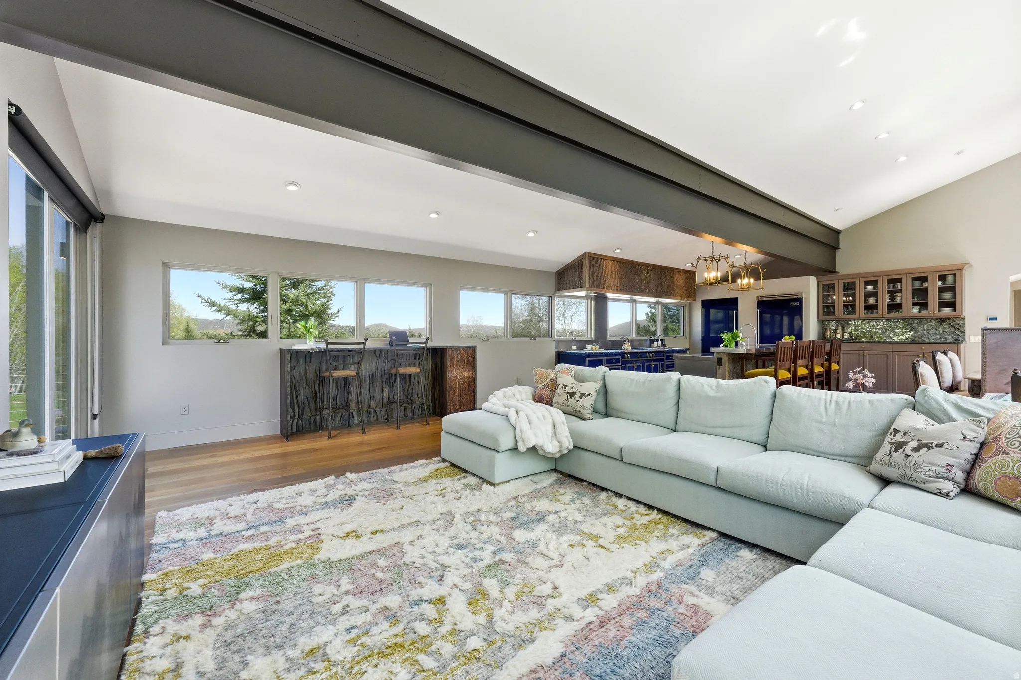 Open-concept living space featuring wood-finish flooring, a vaulted ceiling with exposed beam, recessed lighting, a built-in bar area with stone-finish countertop, and a built-in display cabinet