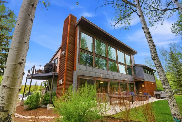 Contemporary property with extensive window fenestration, a sizable upper-level deck with metal railings, a ground-level patio, and a mix of vertical wood-finish siding and horizontal paneling