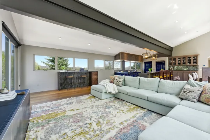 Open-concept living space featuring wood-finish flooring, a vaulted ceiling with exposed beam, recessed lighting, a built-in bar area with stone-finish countertop, and a built-in display cabinet
