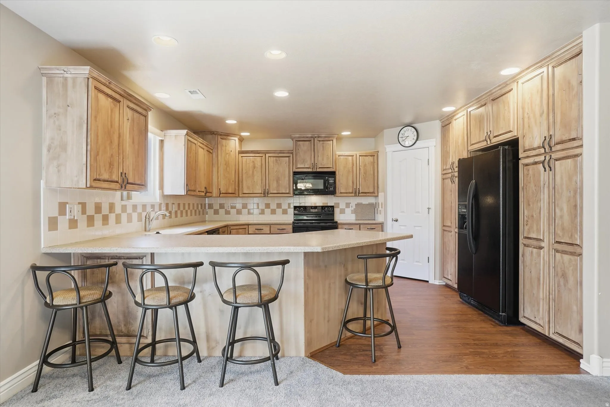 Kitchen featuring wood-finish cabinetry, a large peninsula with countertop seating, a tile backsplash, recessed lighting, and black appliances