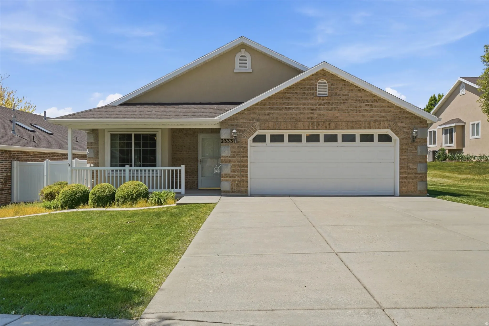 Brick facade home featuring a two-car garage with transom windows, covered front porch with white railing, and a manicured lawn with mature shrubs