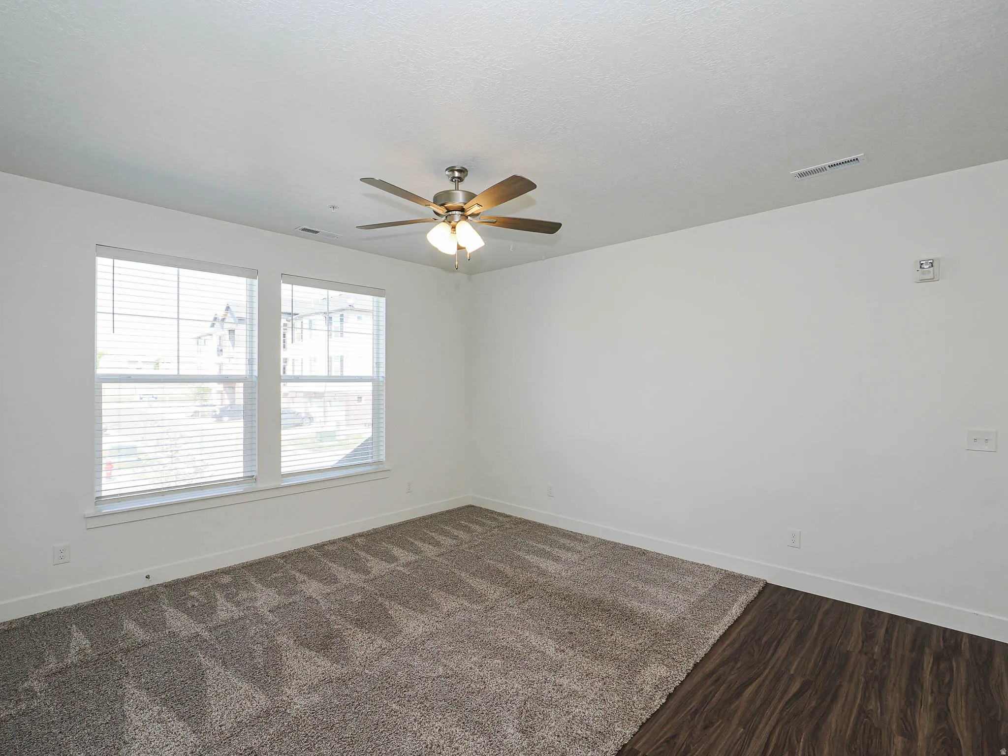 Room featuring two large windows with horizontal blinds, a ceiling fan with integrated lighting, wood-finish flooring, and wall-to-wall carpeting