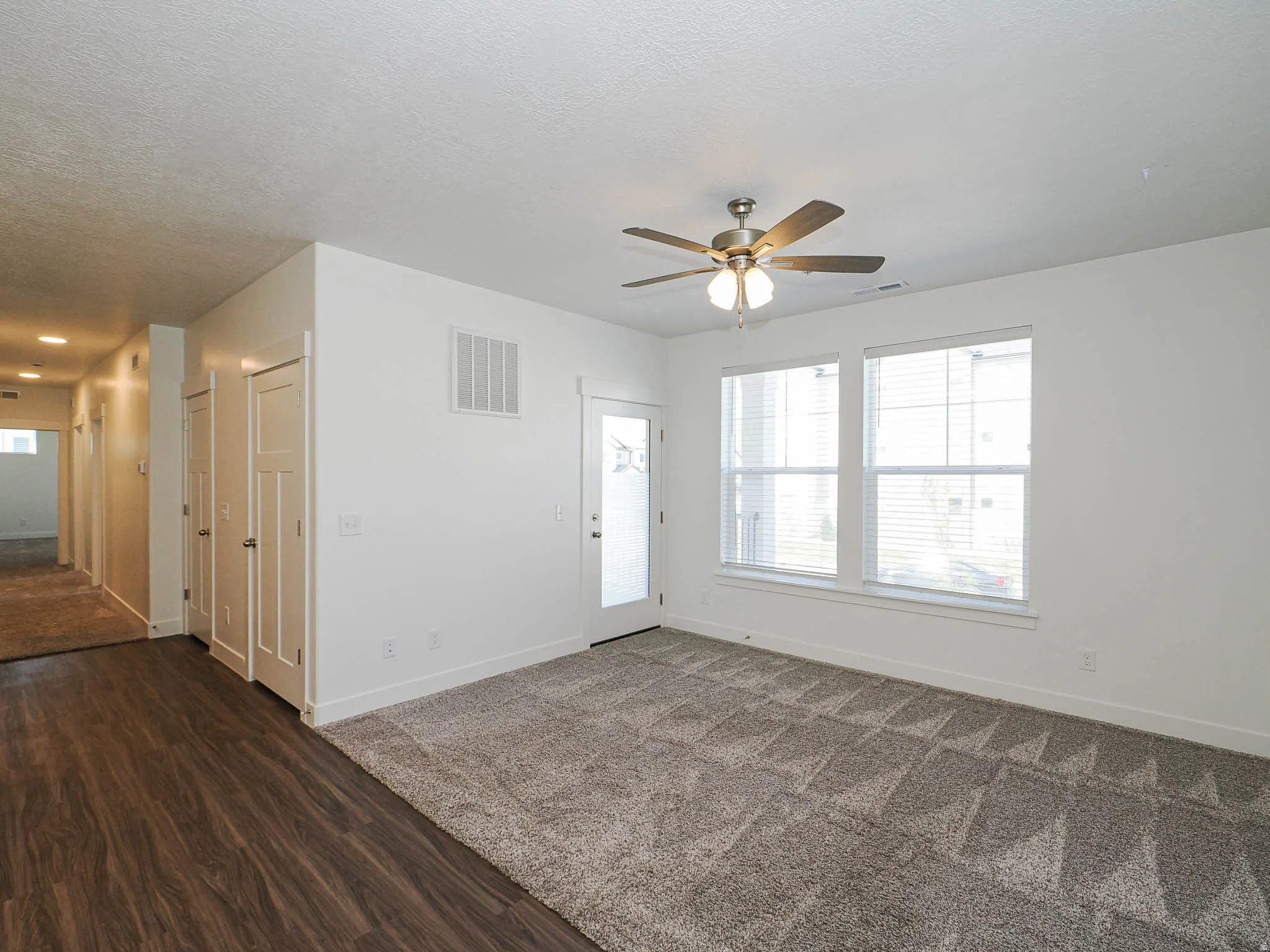 Spacious room featuring neutral carpeting, a ceiling fan with integrated lighting, two large windows with blinds, a glass-paneled door, and recessed lighting in the hallway