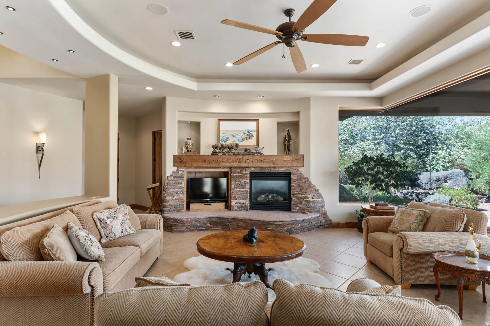 Spacious living area featuring a stone fireplace with a wood-beam mantel, recessed lighting, ceiling fan, tile flooring, and large window overlooking natural scenery