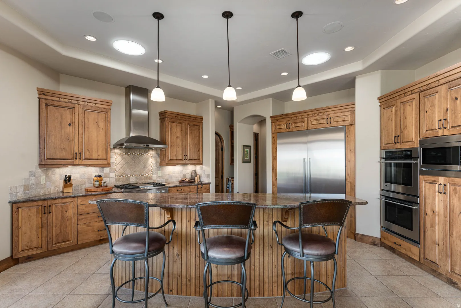 Spacious kitchen featuring an expansive central island with stone-finish countertop, custom wood cabinetry, stainless steel appliances, a tiled backsplash, and recessed lighting with skylights