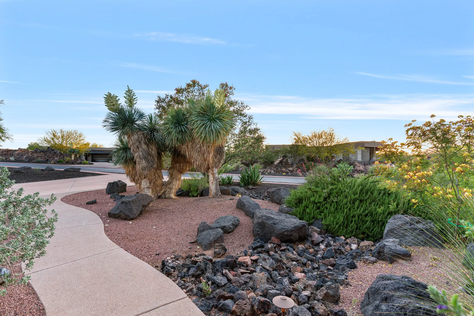 Curved concrete walkway bordered by a xeriscaped garden featuring mature yucca plants, dark volcanic rock, and a red gravel groundcover
