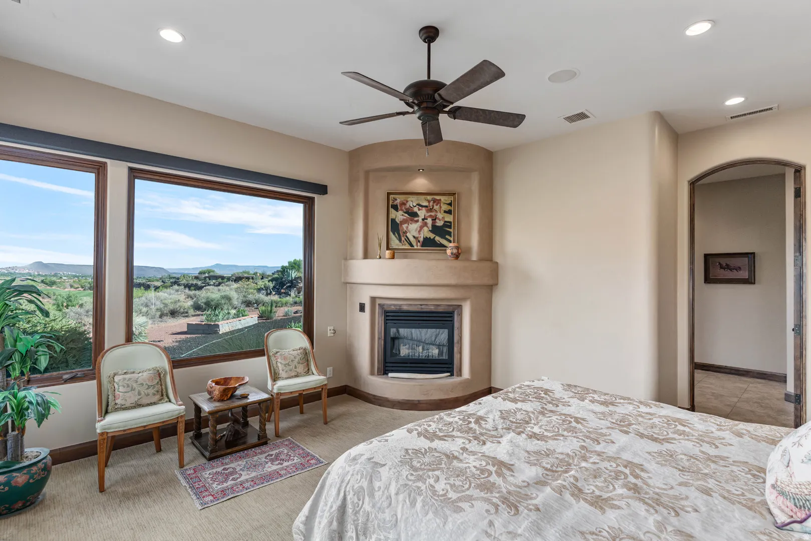 Primary bedroom featuring large windows with a desert landscape, a corner fireplace with a built-in mantel, a ceiling fan, recessed lighting, and an arched doorway