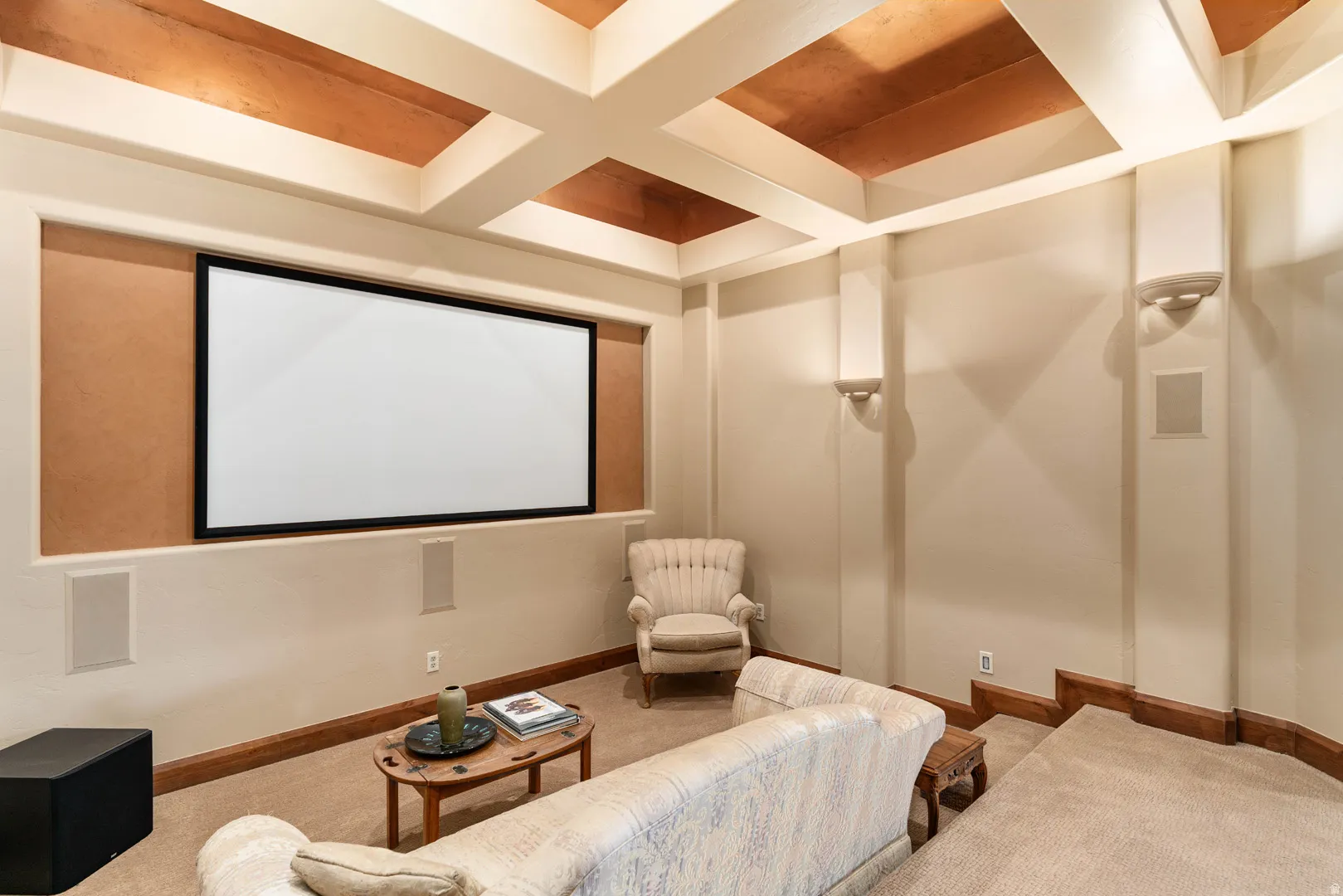 Built-in projection screen with recessed speakers, coffered ceiling with copper-toned panels, wall sconce lighting, and wood-trim baseboards