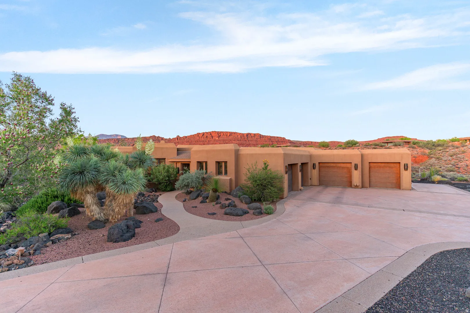 Desert-style exterior featuring a stucco finish, attached three-car garage with wood-finish doors, a winding concrete driveway, rock landscaping, and mature desert plantings