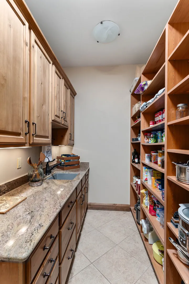 Spacious pantry featuring extensive wood cabinetry, granite countertops, an undermount sink with a dark-finish faucet, under-cabinet lighting, and built-in wood shelving
