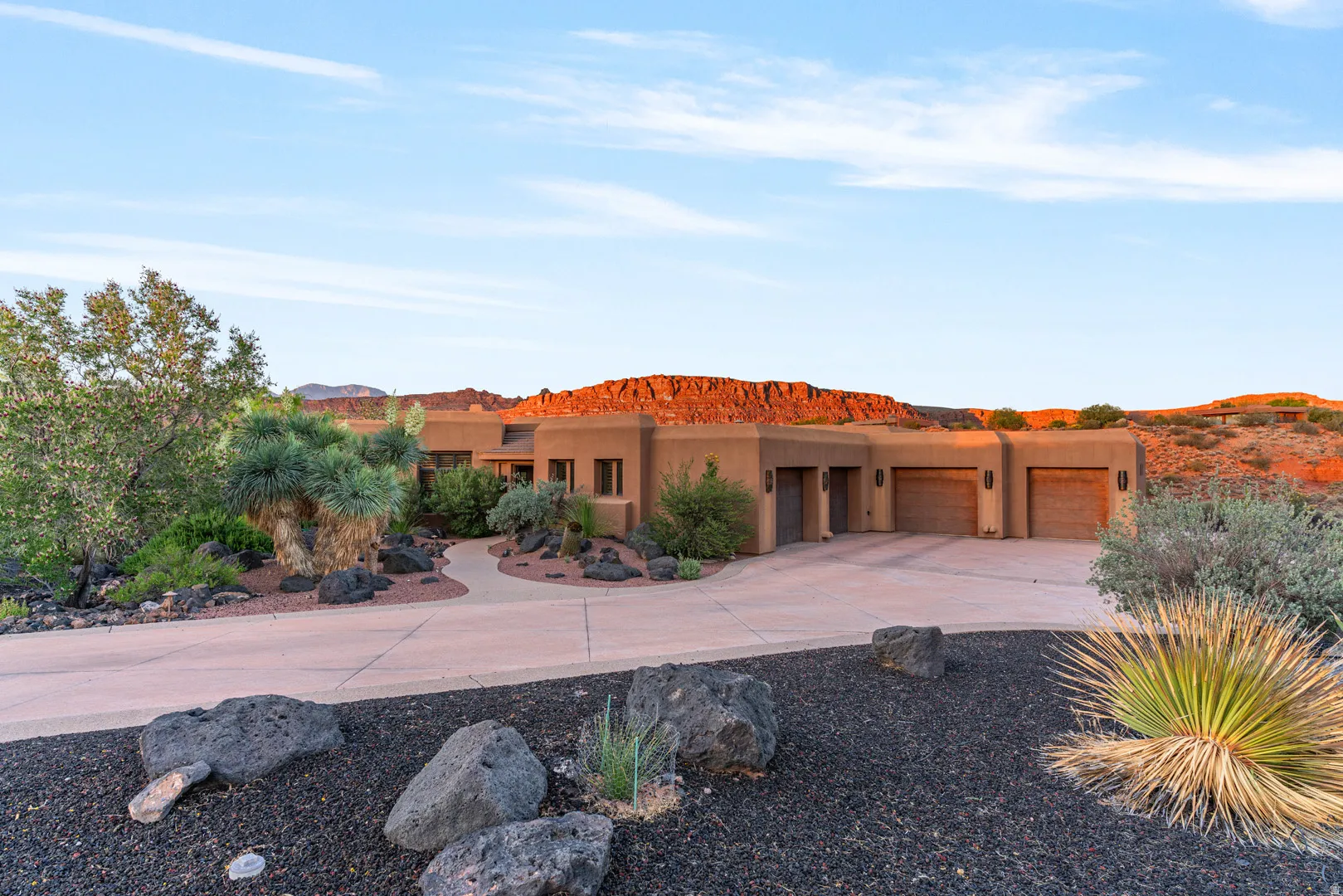 Southwestern style residence featuring a stucco exterior, multiple garage bays with wooden doors, a curving concrete driveway, and desert landscaping with dark volcanic rock accents