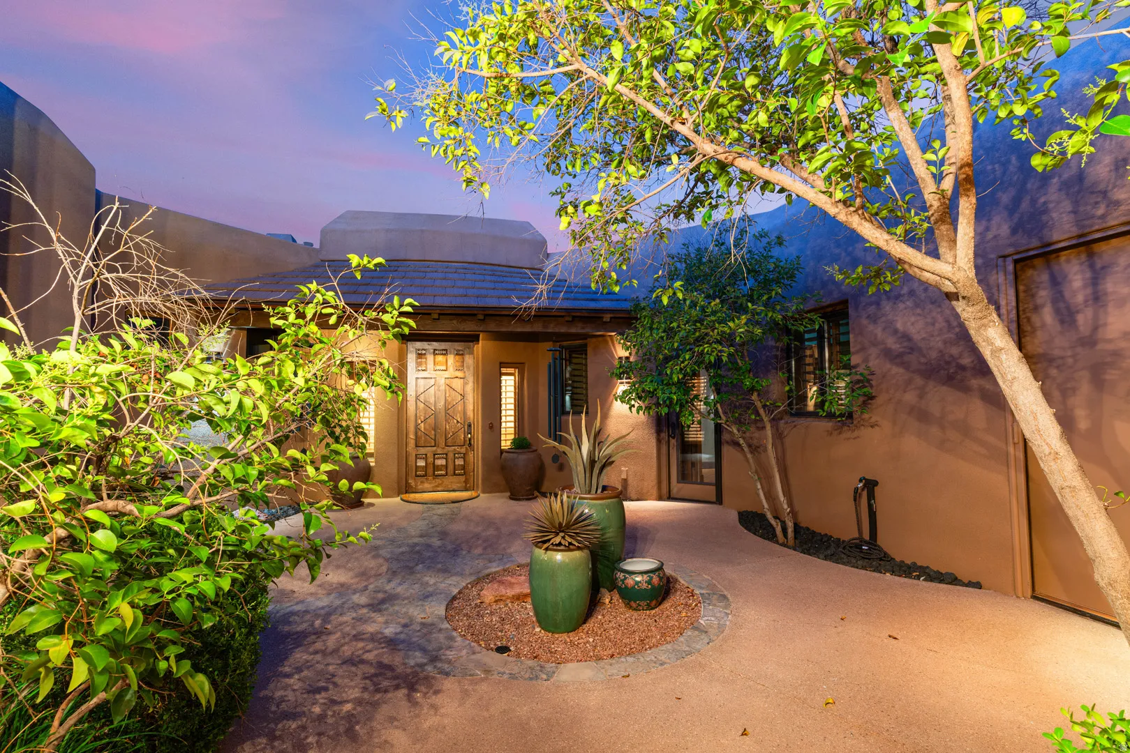Southwestern-style entry courtyard featuring a decorative wooden door, mature landscaping, integrated lighting, and a circular planter with accent stones