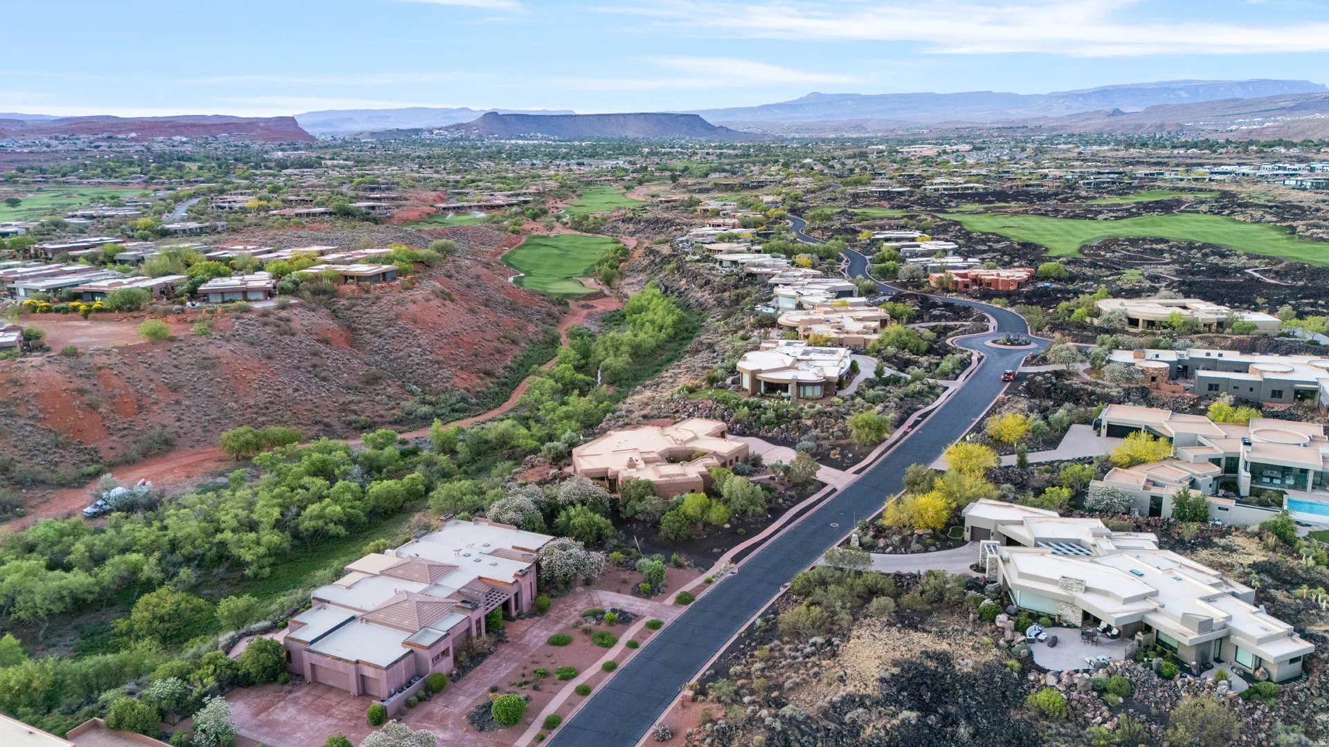 Expansive aerial captures a residential development with varied architectural styles, a paved street, natural desert landscaping, and a golf course in the distance