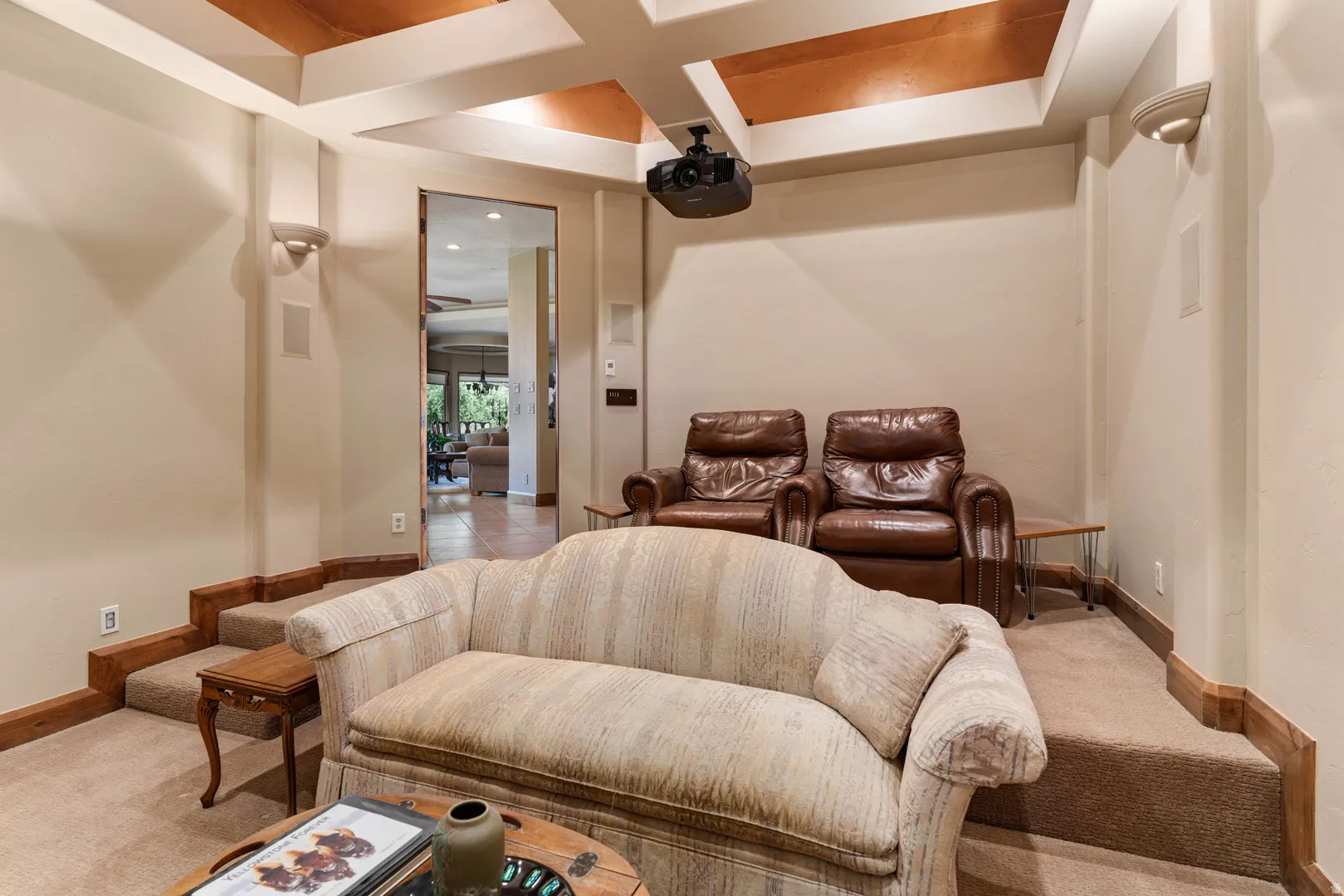 Dedicated media room featuring tiered carpeted flooring, a ceiling-mounted projector, wall-mounted speakers, and decorative coffered ceiling detail with wood-finish panels