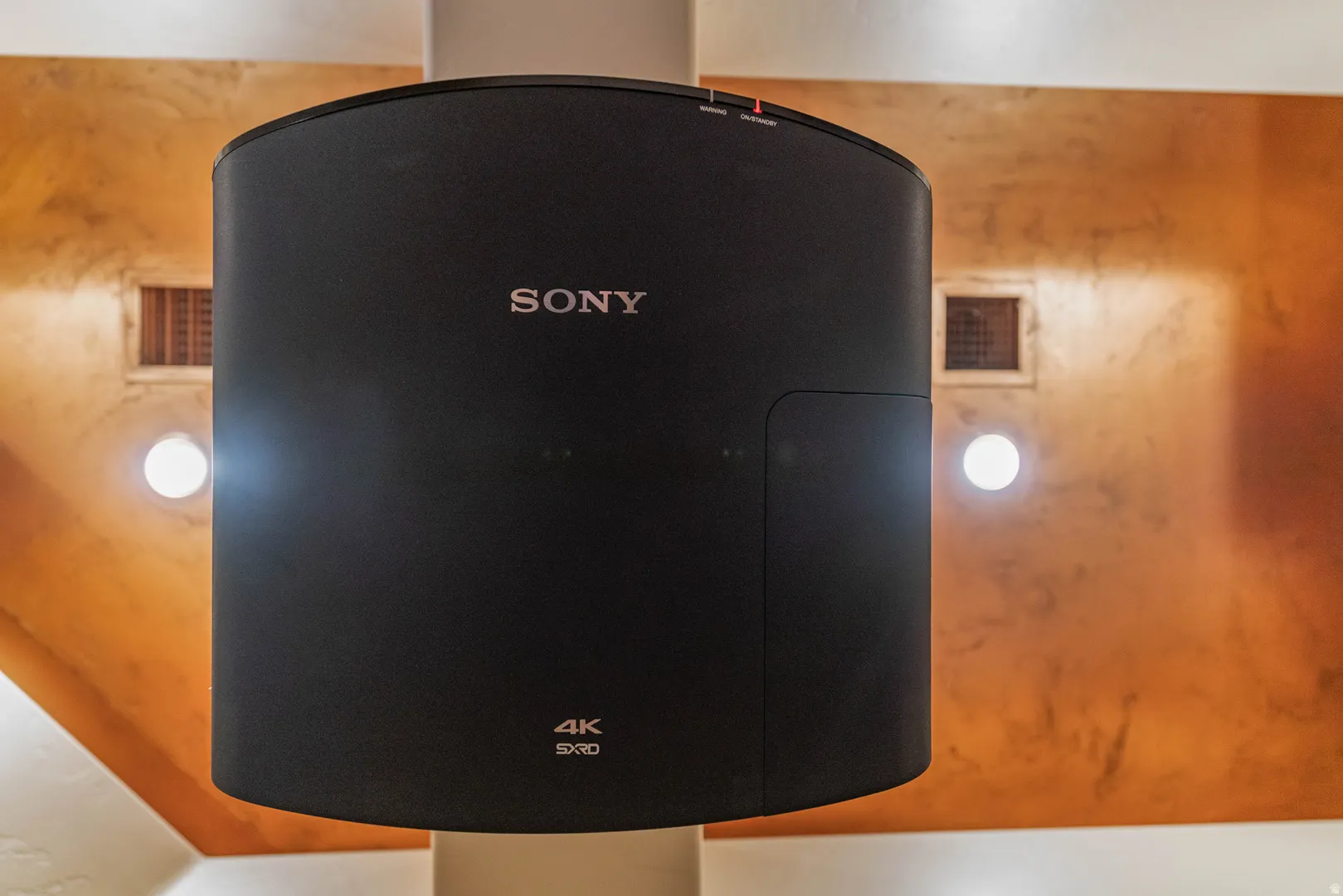 Ceiling-mounted 4K projector with a matte black finish, accented by an orange-hued textured wall, recessed lighting, and a square vent