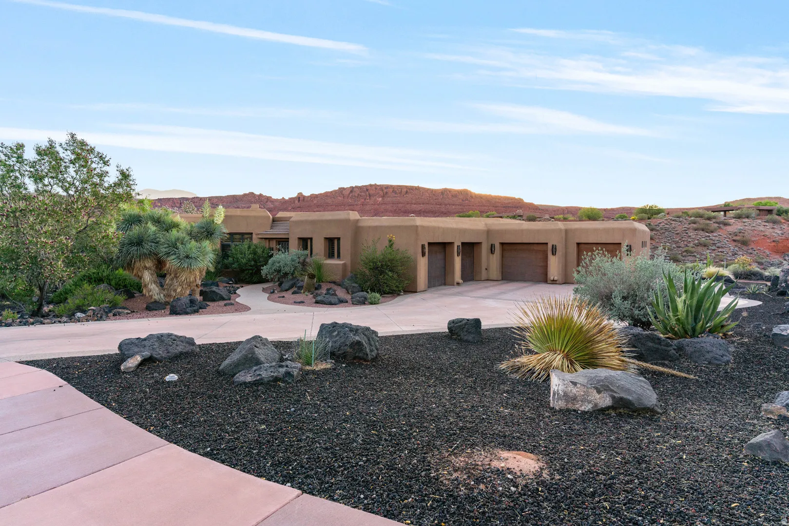 Southwestern stucco exterior with an integrated three-car garage, concrete driveway, and desert landscaping featuring various mature plants and dark rock ground cover