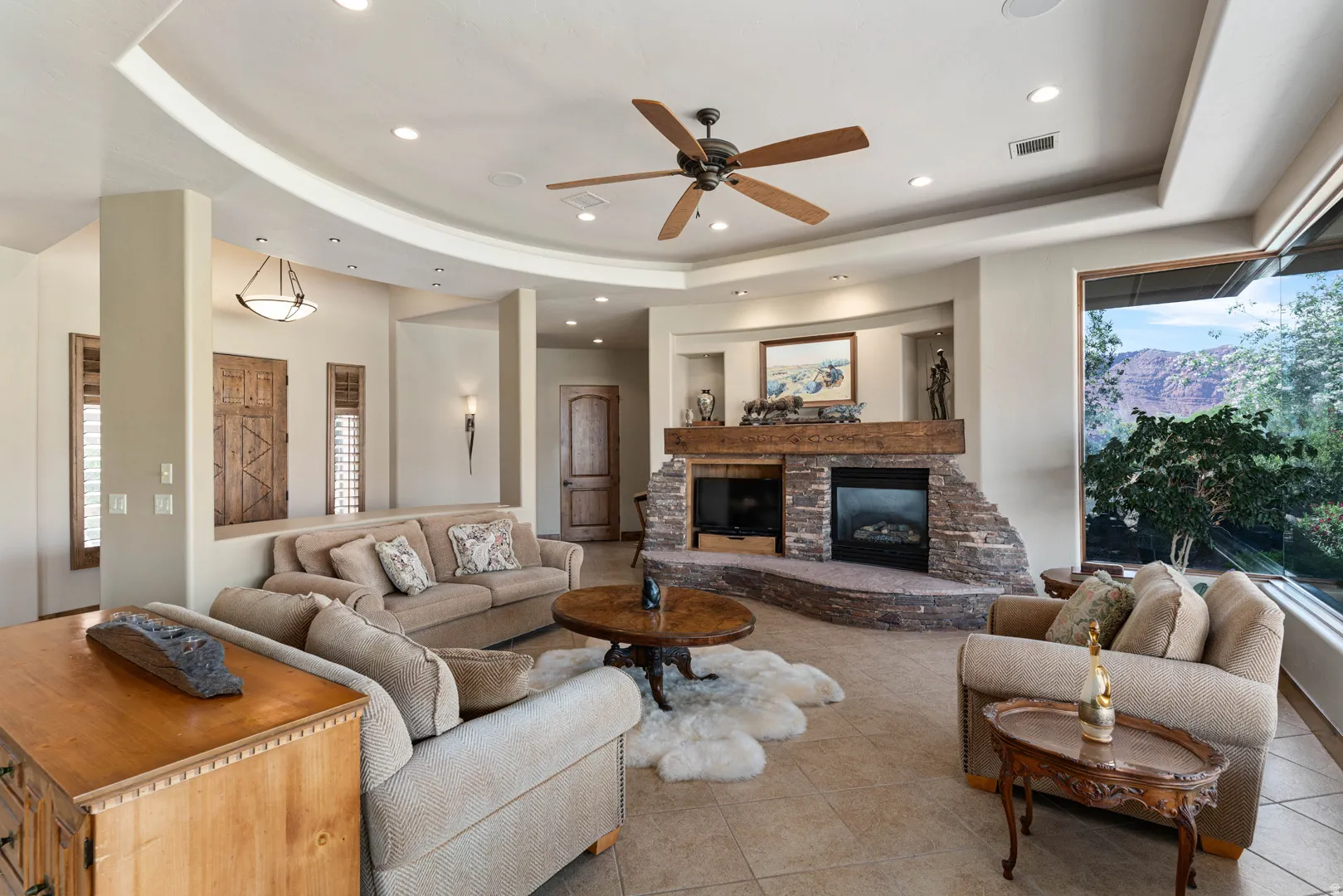 Living area featuring a stone fireplace with a wood mantle, a large picture window, and tray ceilings with recessed lighting and a ceiling fan