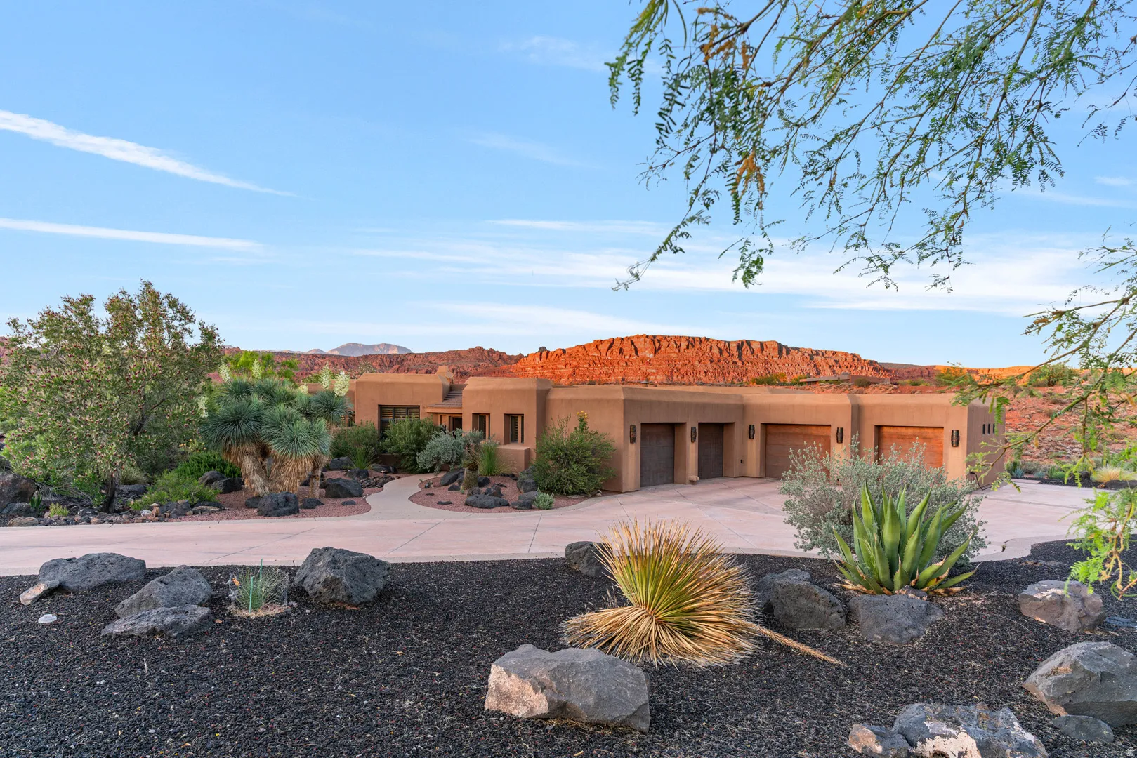 Adobe-style exterior with a paved circular drive, three garage bays featuring wood-finish doors, and natural rock landscaping