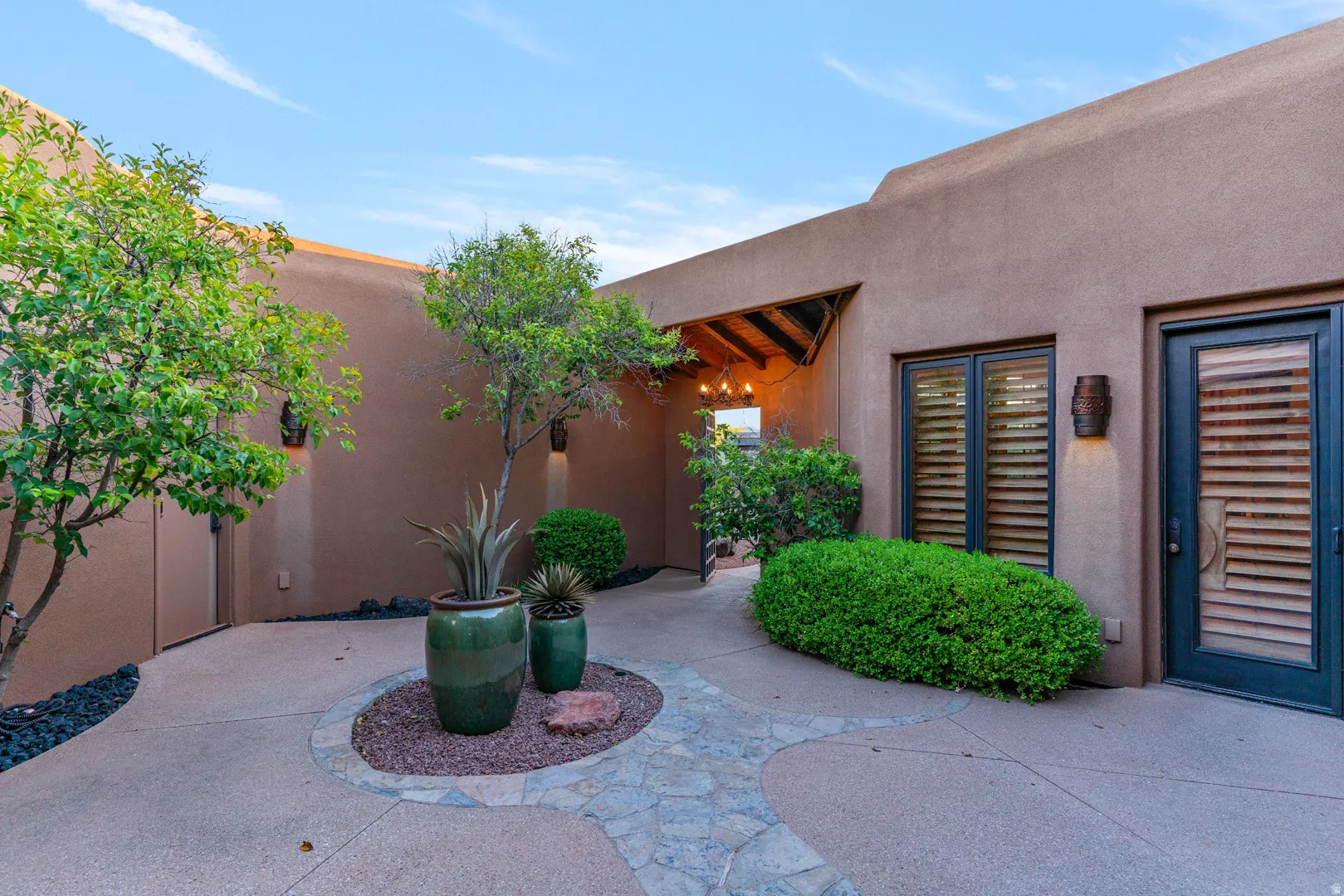Exterior courtyard featuring a flagstone and concrete pathway, mature trees, and integrated stucco walls with sconce lighting