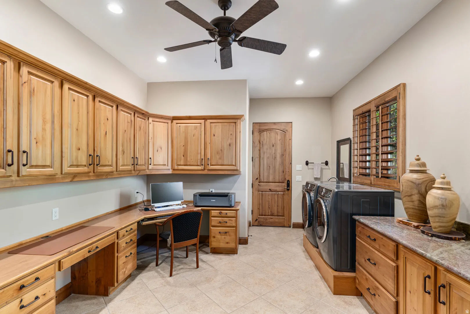 Spacious utility room featuring extensive natural wood cabinetry, a built-in desk, tile flooring, a dark-finish ceiling fan, and recessed lighting