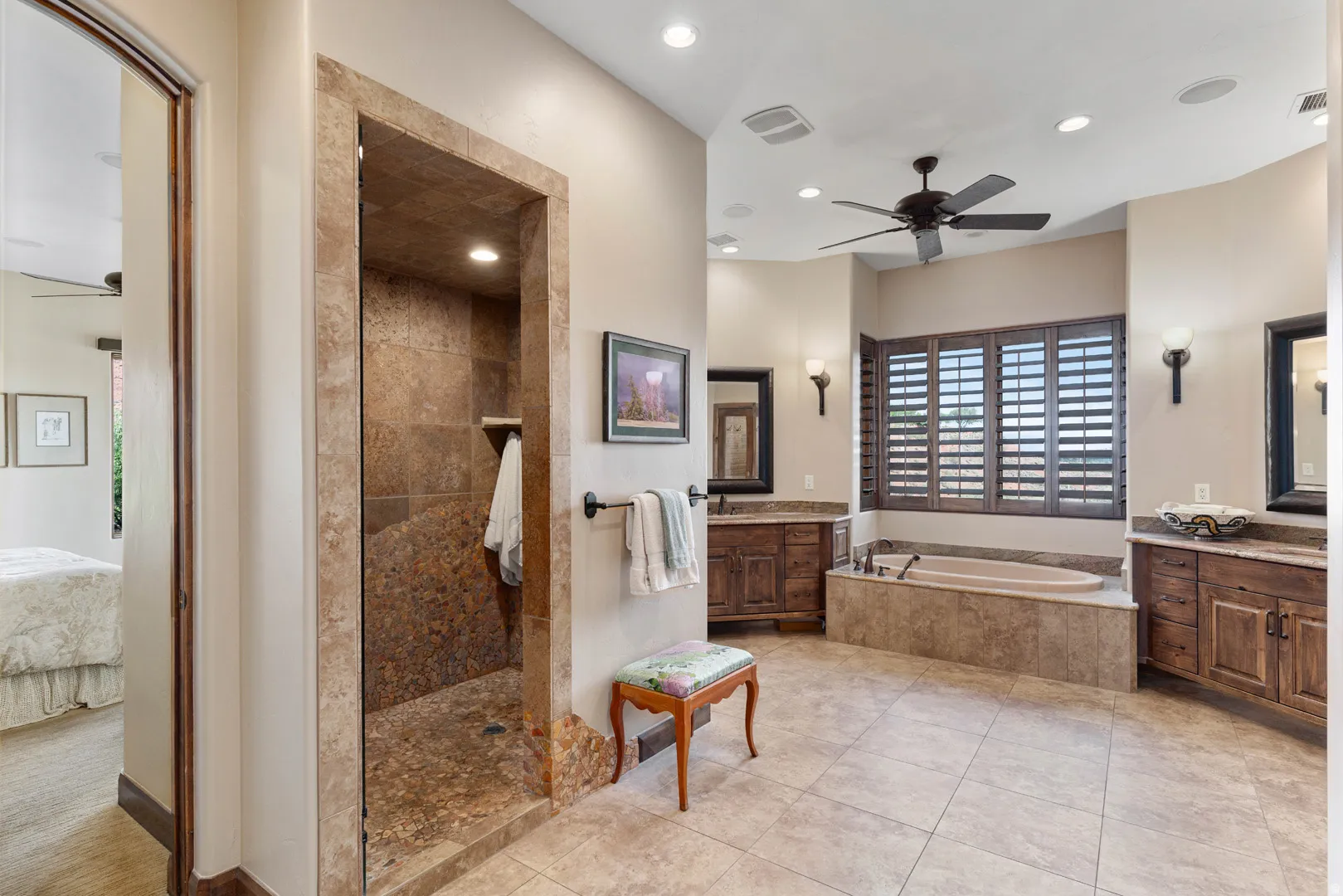 Spacious bathroom featuring a large soaking tub with a tiled surround, a walk-in shower with stone-tiled walls and pebble flooring, dual vanity areas with vessel sinks, and a ceiling fan