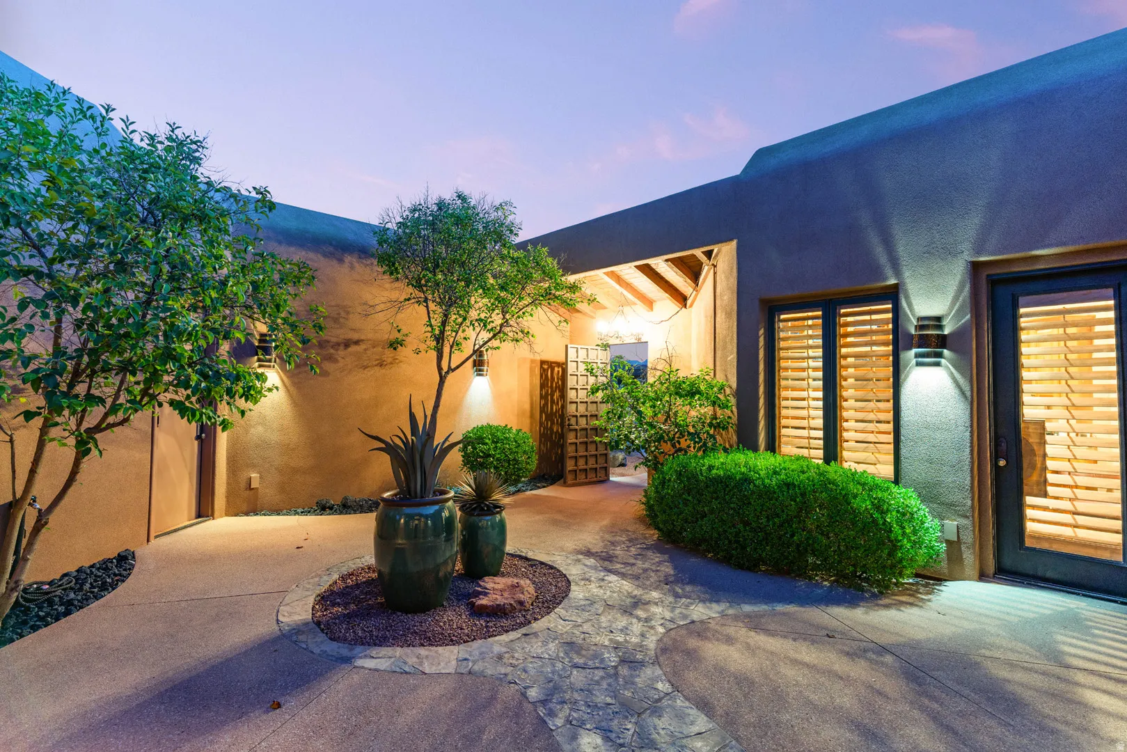 Entry courtyard featuring a stone pathway, stucco exterior, built-in wall sconces, natural wood shutters, and mature potted plants