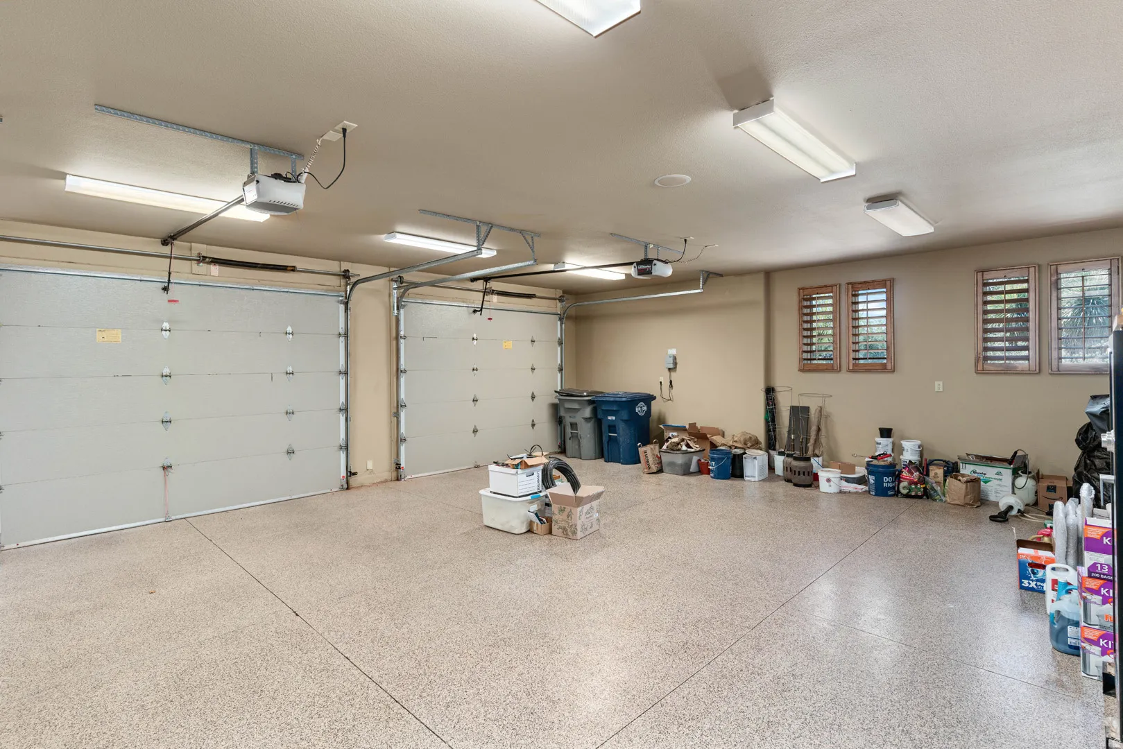 Spacious garage featuring a speckled epoxy floor, two overhead garage doors, multiple fluorescent light fixtures, and three windows with wooden slat blinds