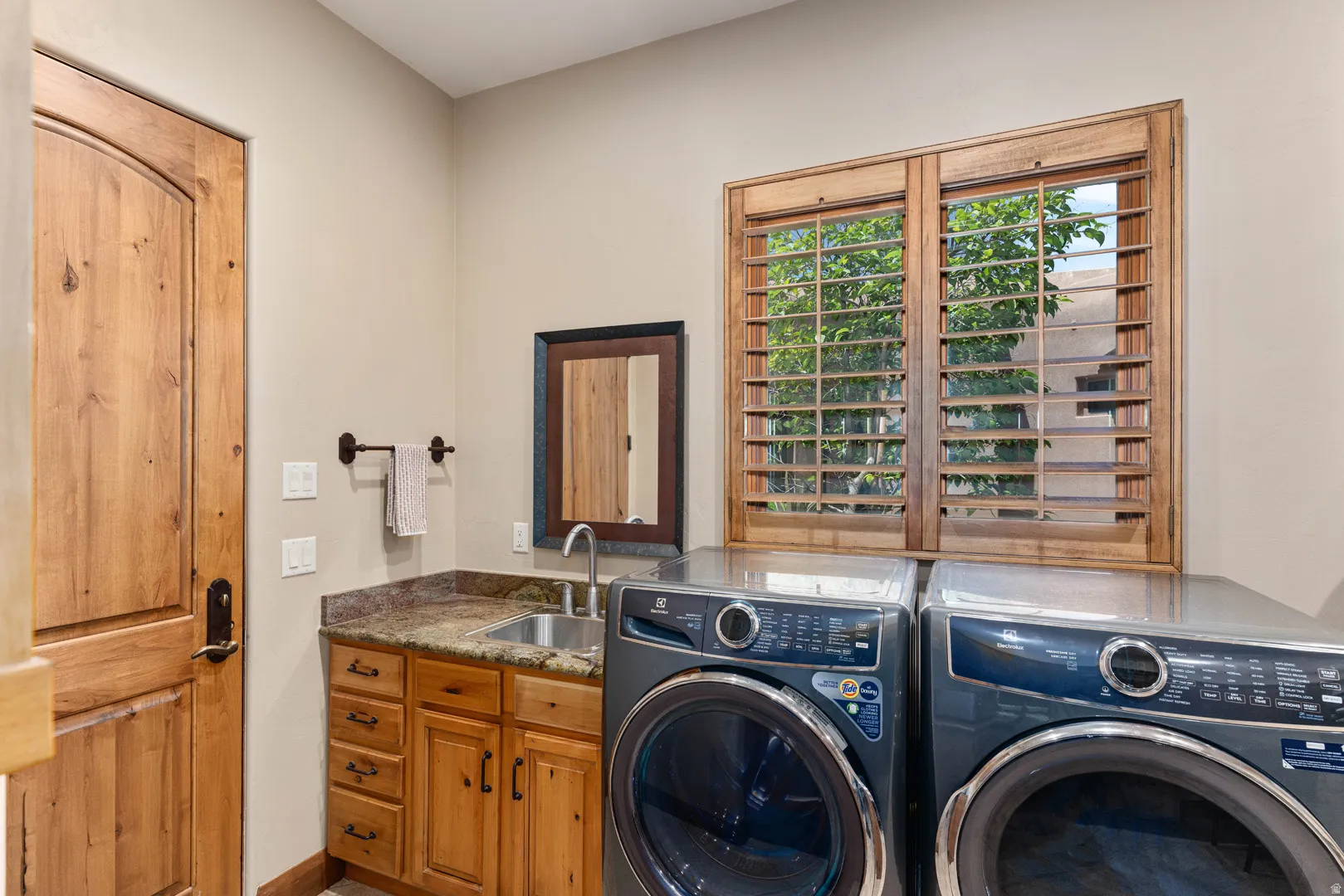 Laundry room featuring a utility sink with a stone-finish countertop, wood-finish cabinetry, and a window with wood shutters
