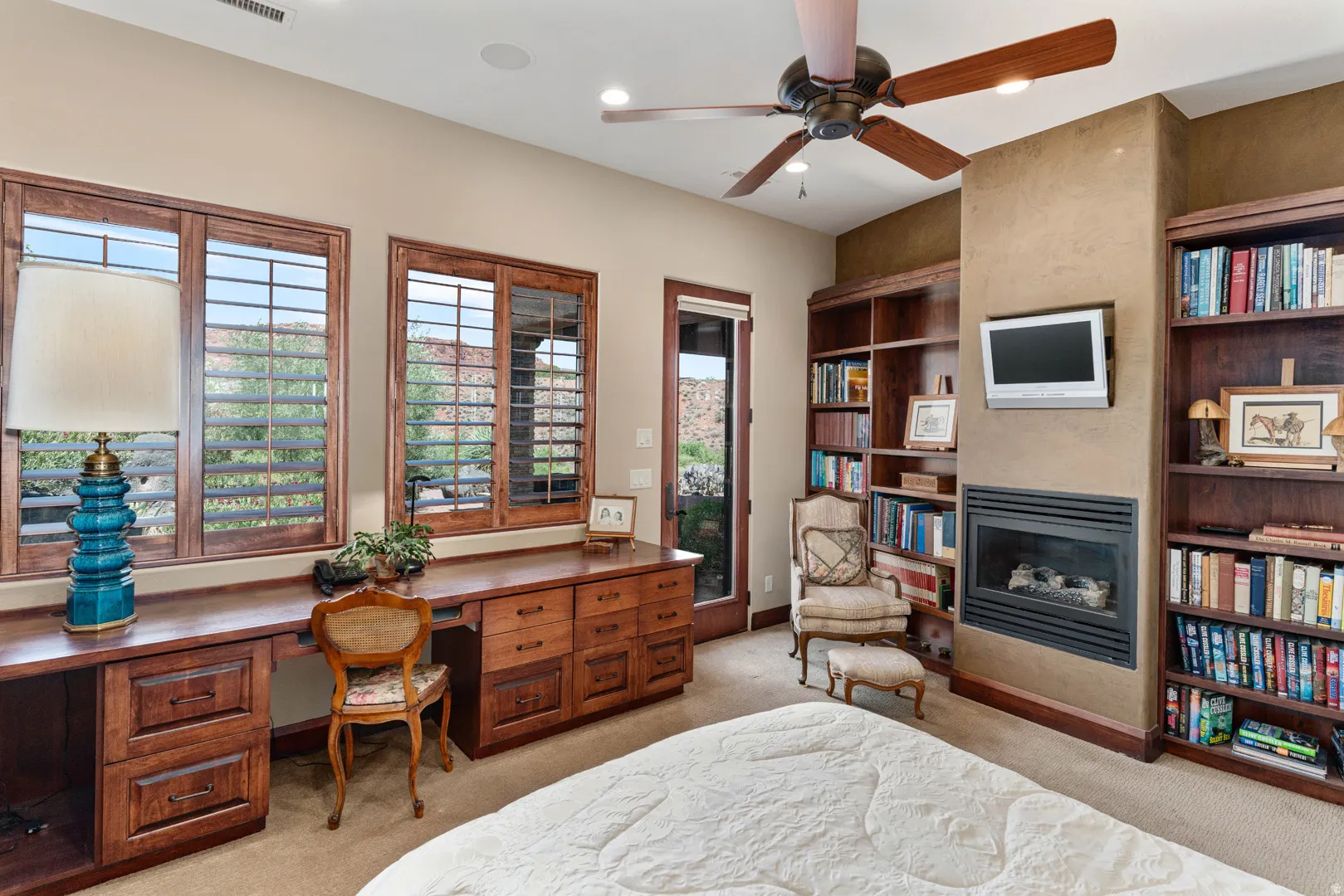 Integrated wood-finish desk with storage cabinetry, two large windows with plantation shutters, a ceiling fan, and a built-in fireplace with flanking bookshelves