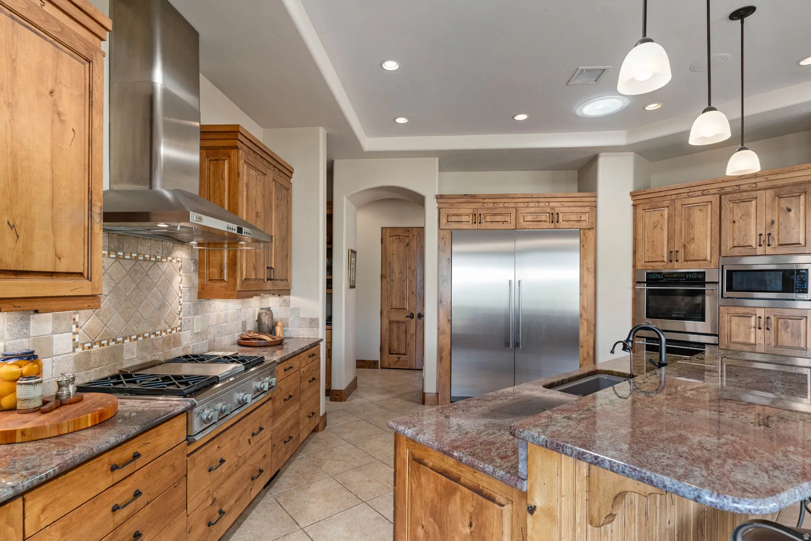 Gourmet kitchen featuring wood cabinetry, stainless steel appliances, granite countertops, a gas range with an overhead exhaust hood, and a tile backsplash