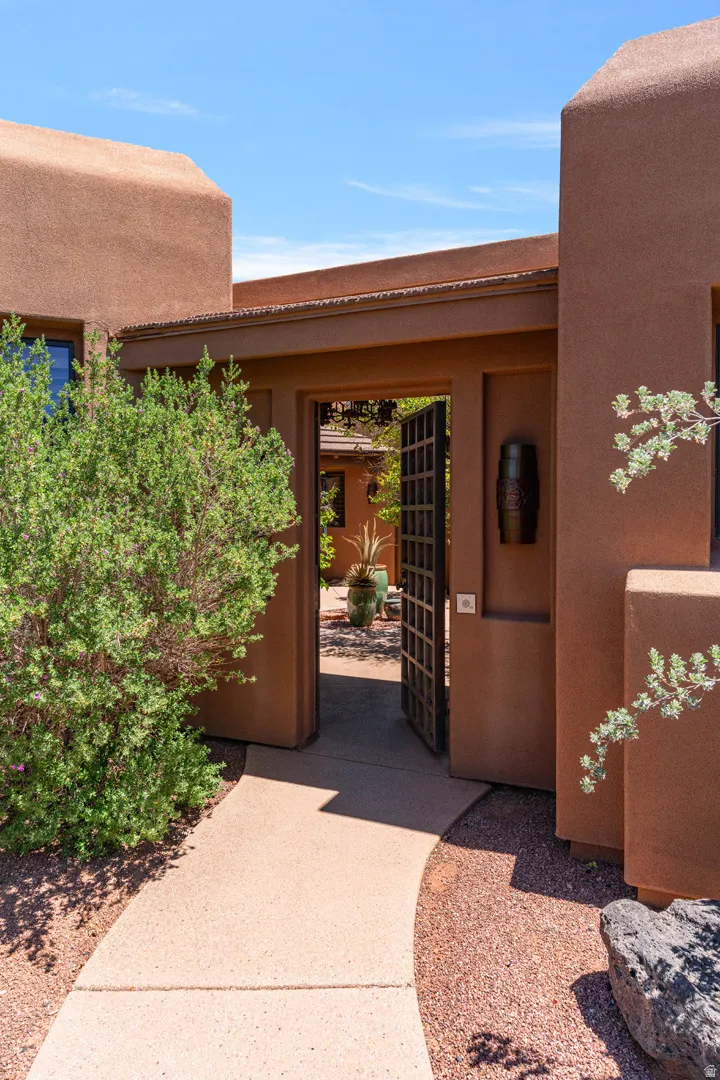 Curved concrete pathway leading to a stucco exterior with an open decorative metal gate, landscaping with mature shrubs and gravel accents