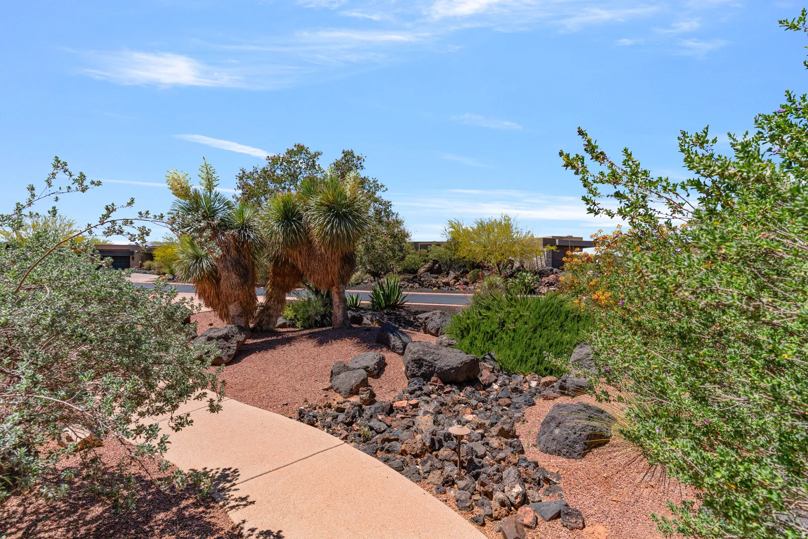 Desert landscaping featuring a stone walkway, mature yucca plants, scattered boulders, and various shrubs