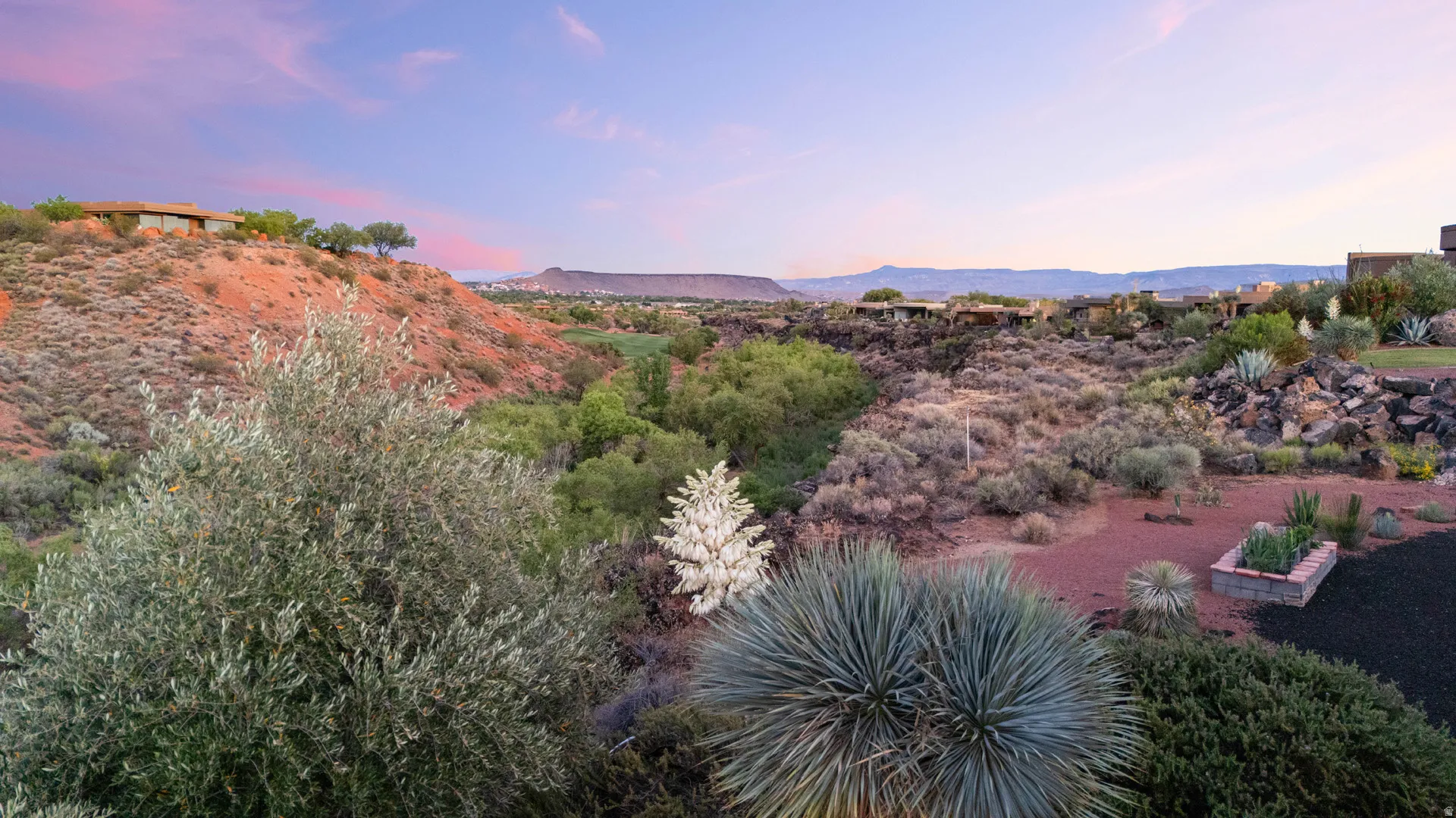 Expansive natural landscape featuring varied desert flora, a rock-lined garden bed, red rock formations, and distant mountain ranges