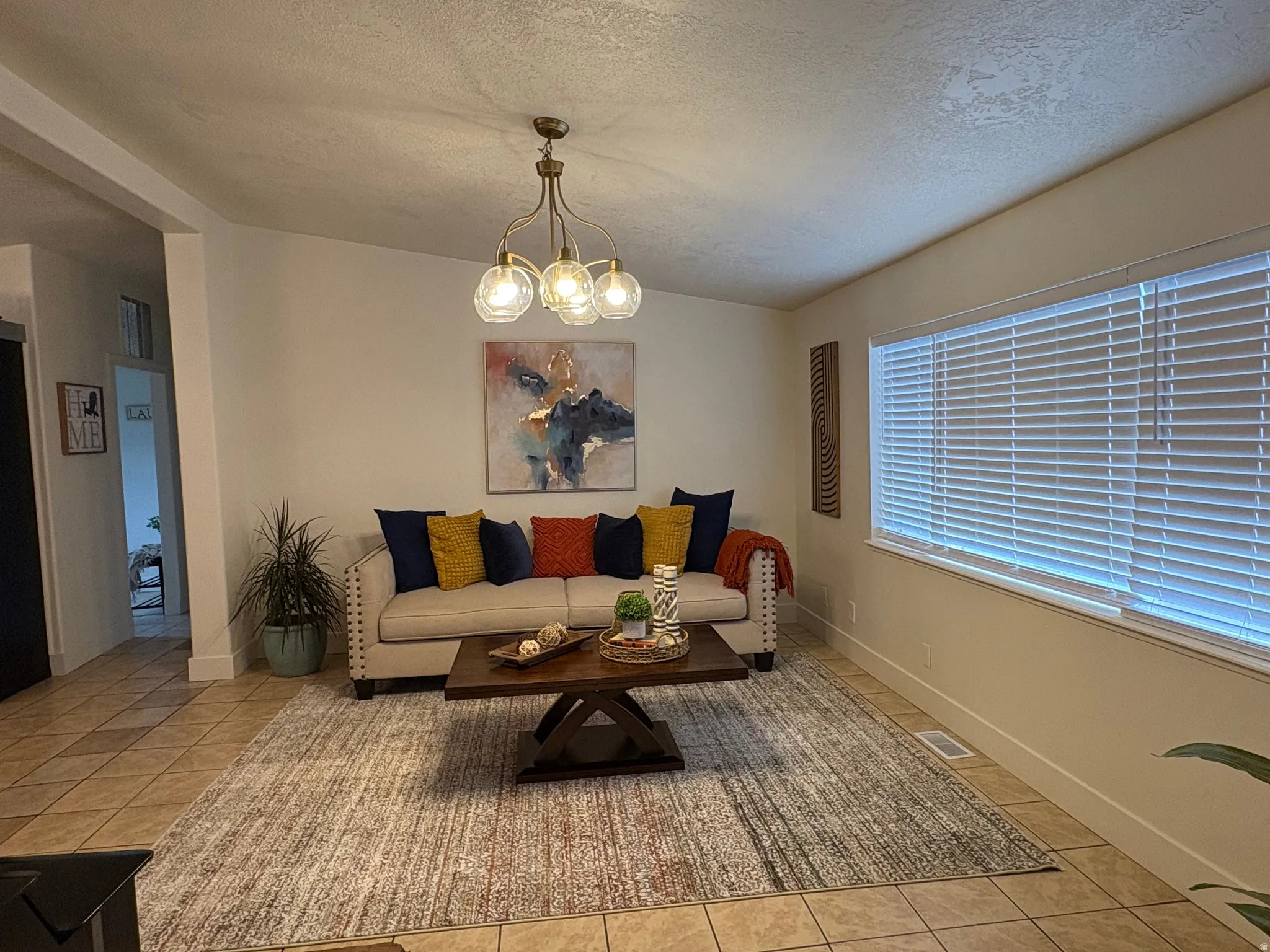 Spacious living area featuring tile flooring, a large window with blinds, and a multi-bulb ceiling fixture