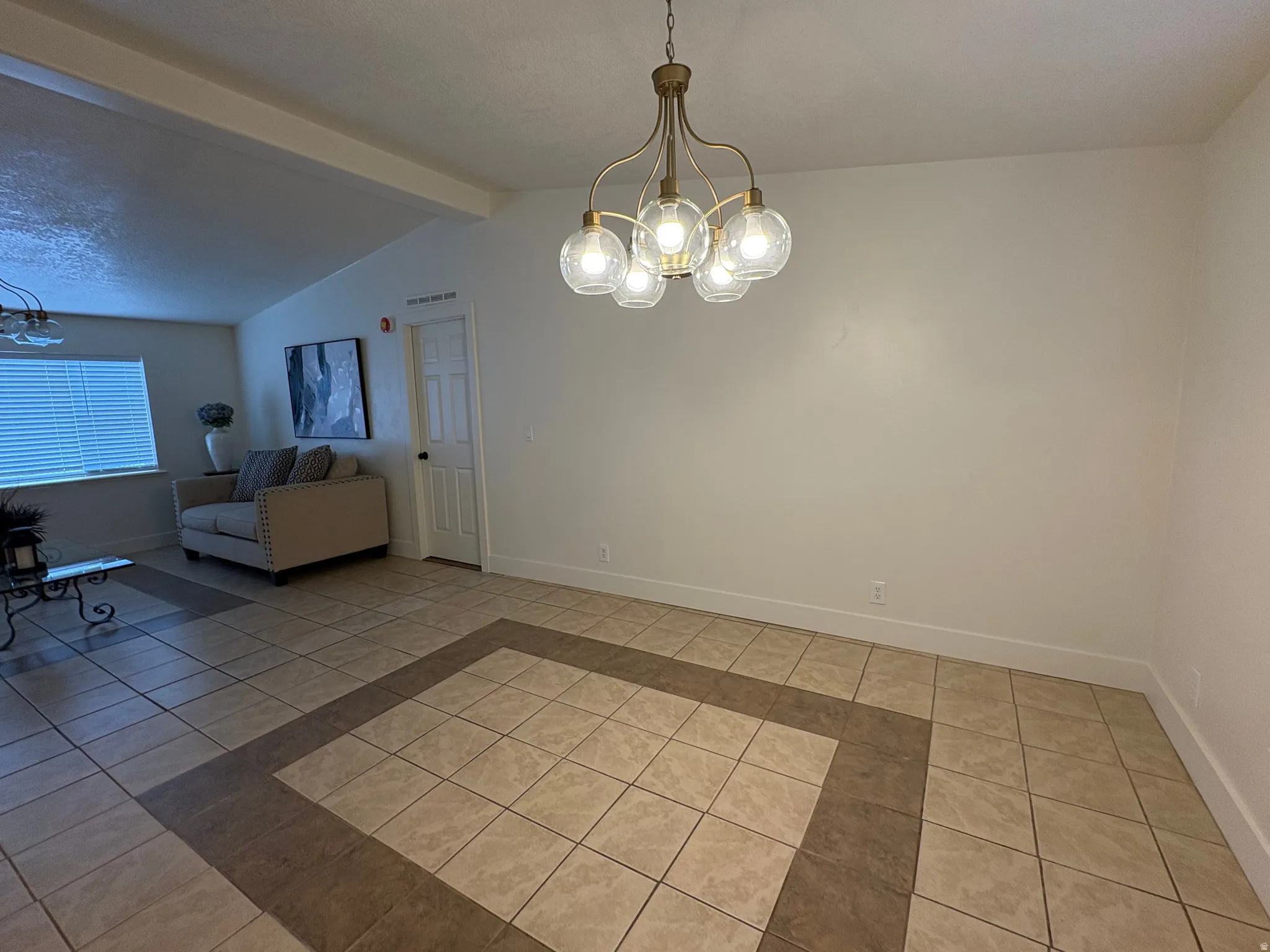 Spacious room featuring a multi-bulb chandelier with clear glass globes, tile flooring with a decorative border, and a window with horizontal blinds