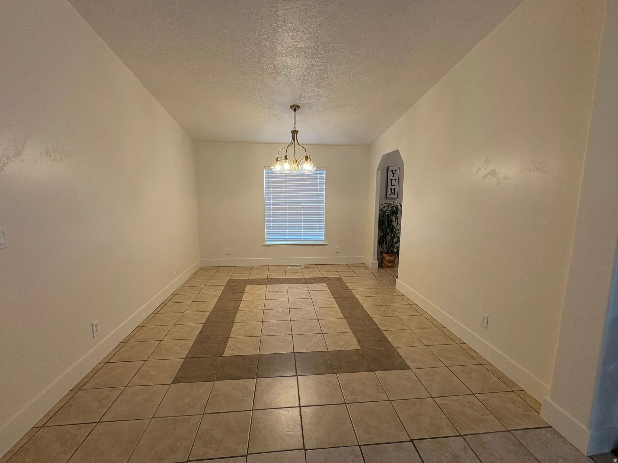 Spacious room featuring neutral-toned tile flooring with a darker accent border, a window with blinds, and an arched doorway