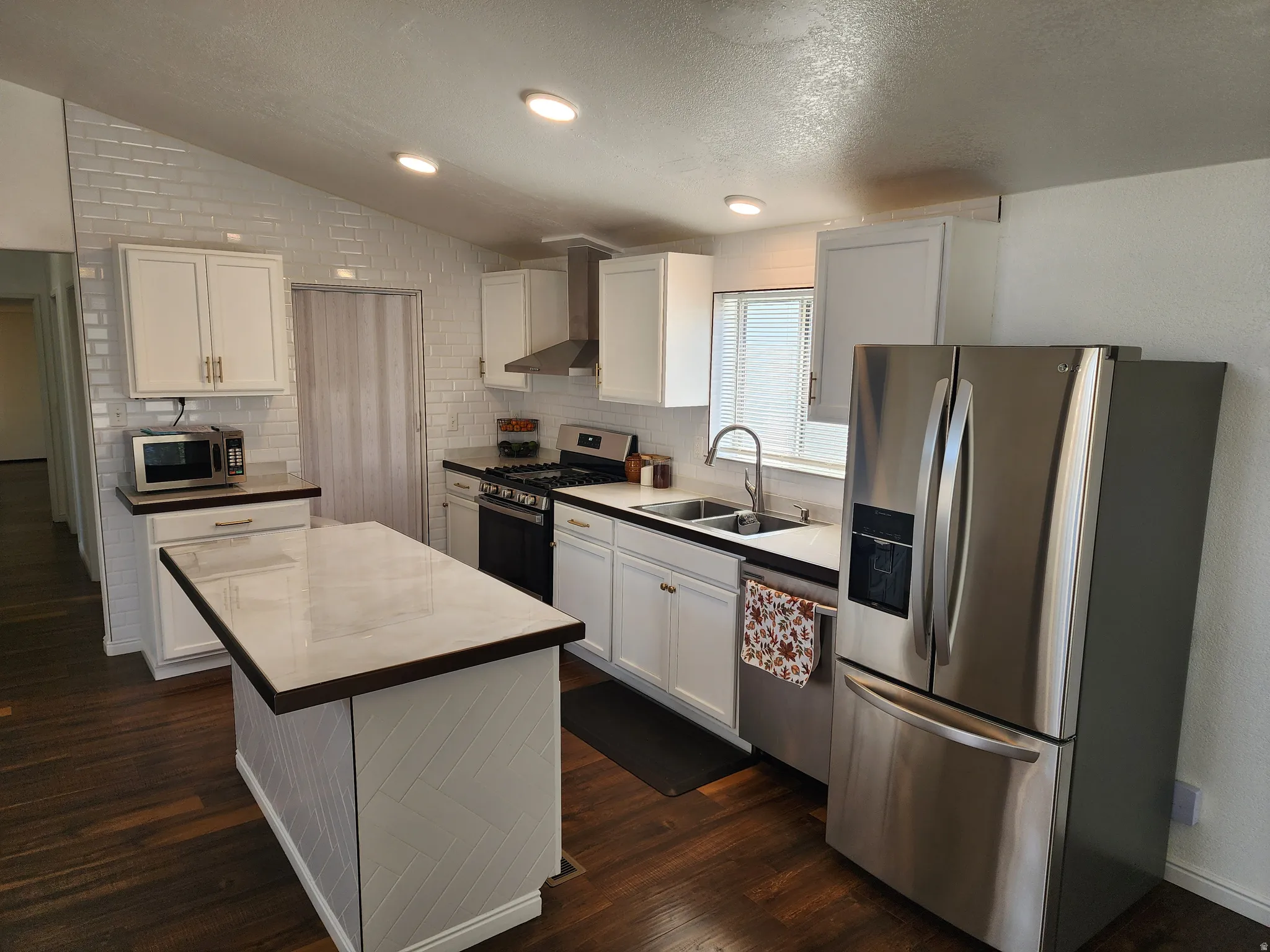 Kitchen featuring a central island with a light-toned countertop, wood-finish flooring, white cabinetry, stainless steel appliances, and a white subway tile backsplash extending to the vaulted ceiling