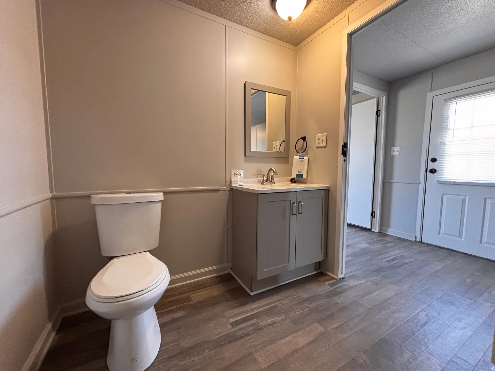Bathroom featuring a white toilet, a gray vanity with a white countertop and integrated sink, a framed mirror, and wood-finish flooring
