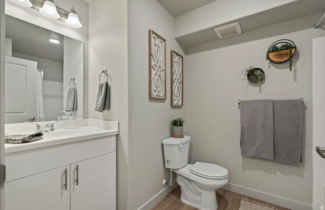 Bathroom featuring a vanity with a white countertop and base cabinetry, a wall-mounted mirror, brushed nickel light fixture, wood-finish flooring, and a white toilet